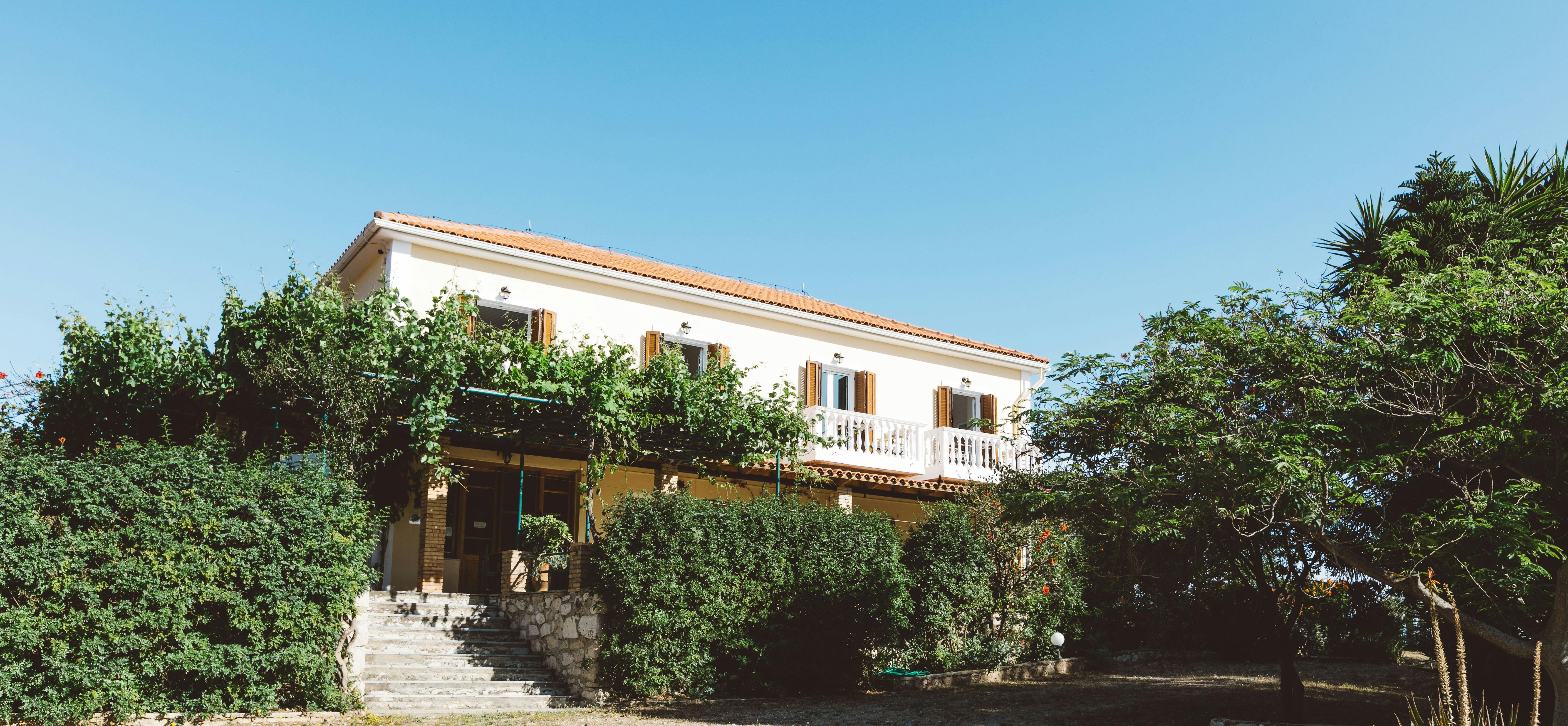 Two-story Mediterranean villa with terracotta roof, white balustrade balcony, and lush garden surroundings under clear blue sky.