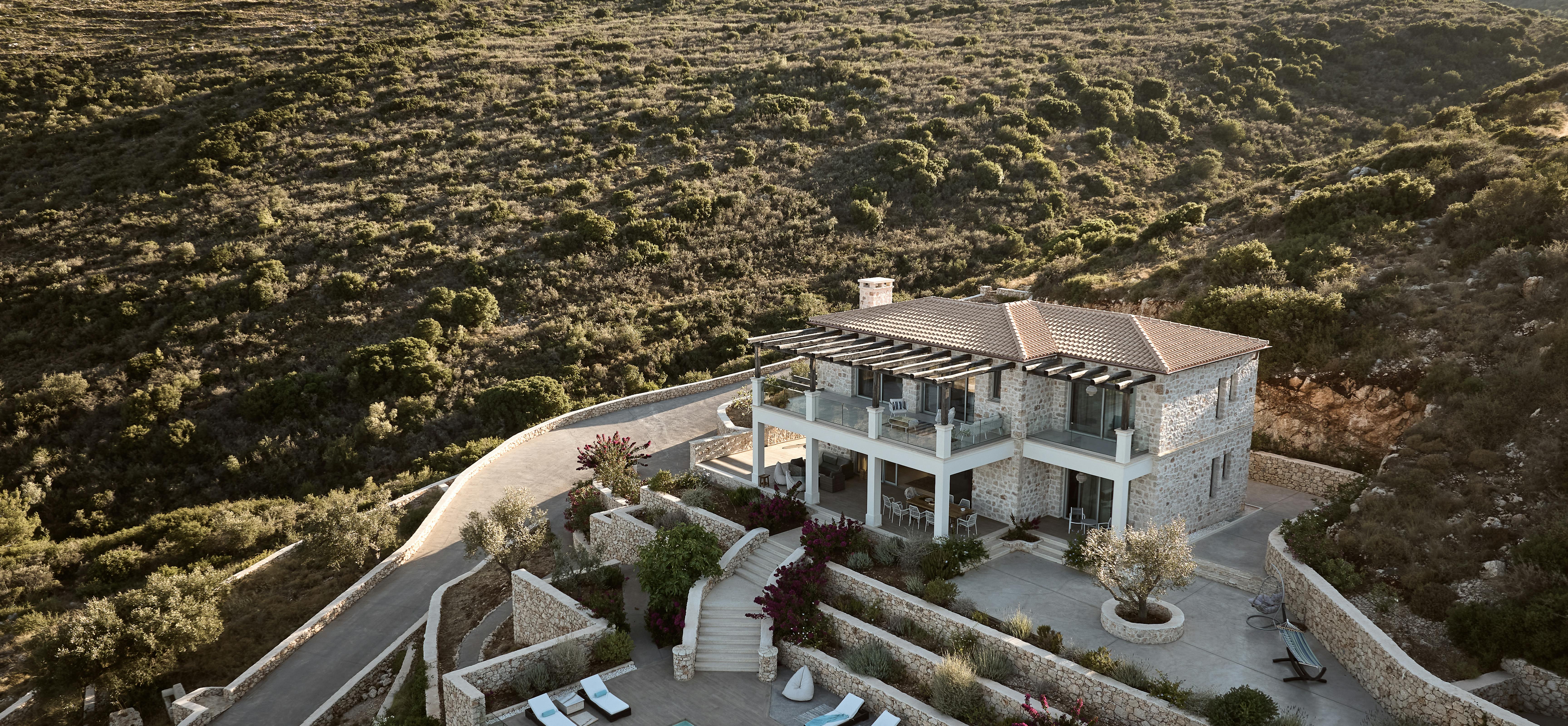 Dramatic aerial perspective of two-story stone villa with curved terracing, infinity pool, multiple outdoor lounging areas, and steep hillside landscape integration.