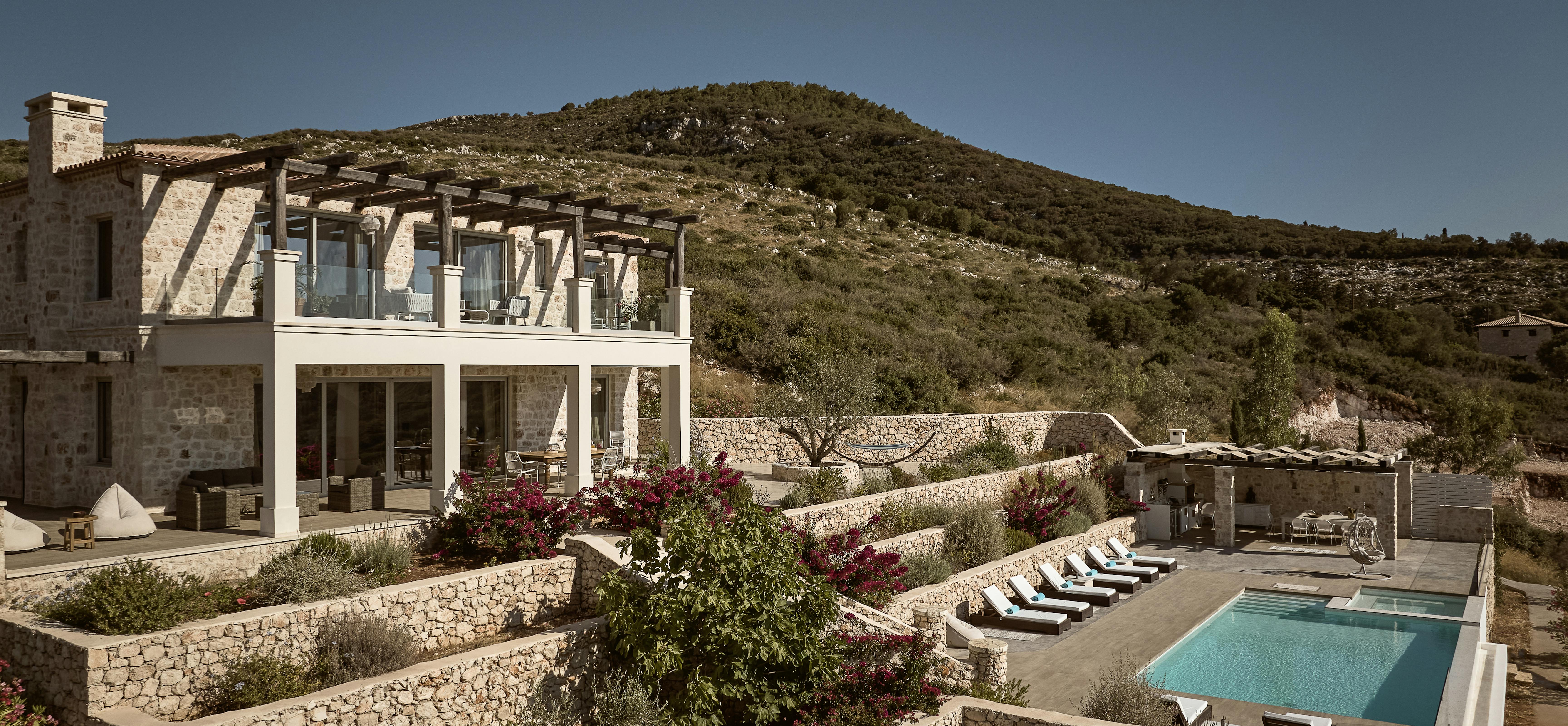 Two-story stone villa with wooden pergola upper terrace, terraced landscaping with native plants, infinity pool, and hillside setting with dramatic elevation.