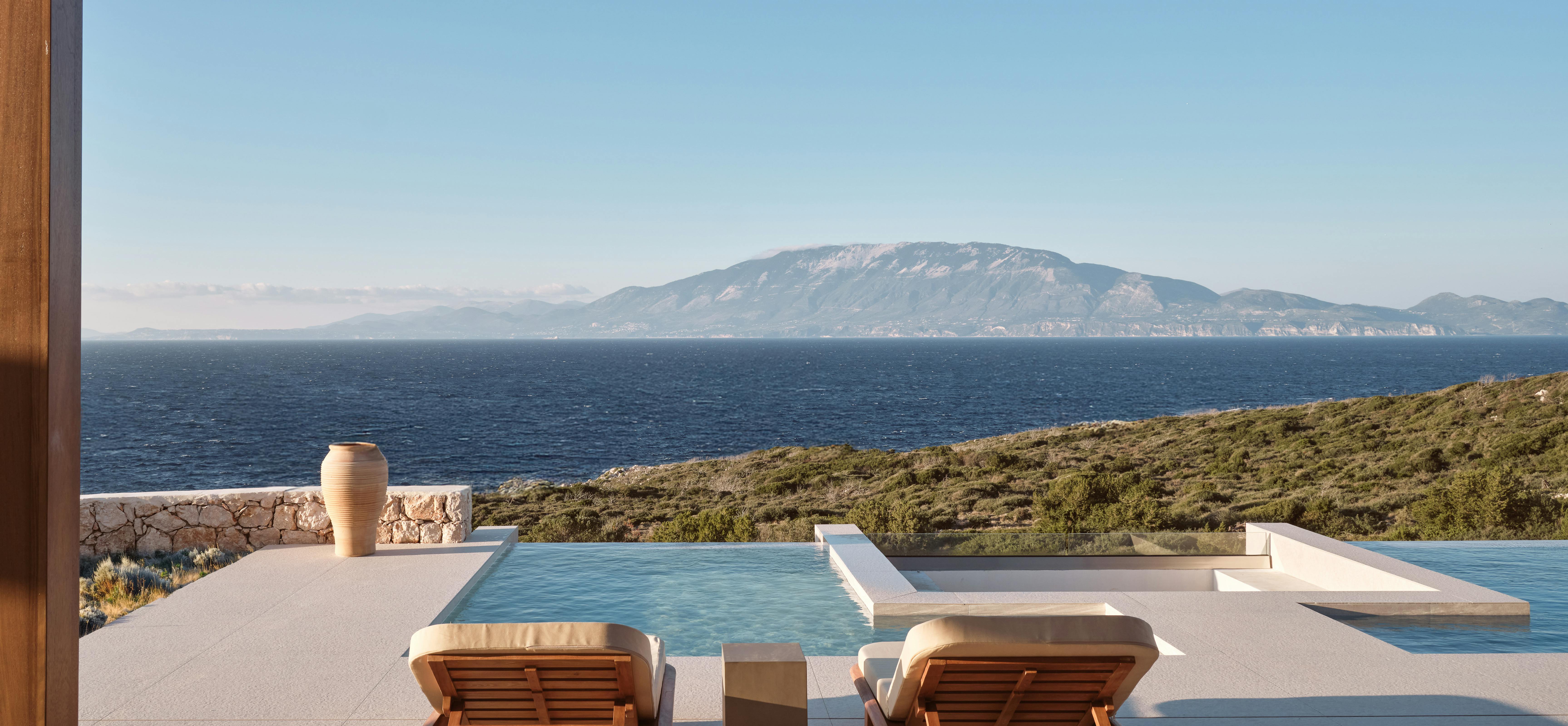 Covered terrace with wooden loungers overlooking an infinity pool and panoramic sea views toward distant mountains under clear blue sky.