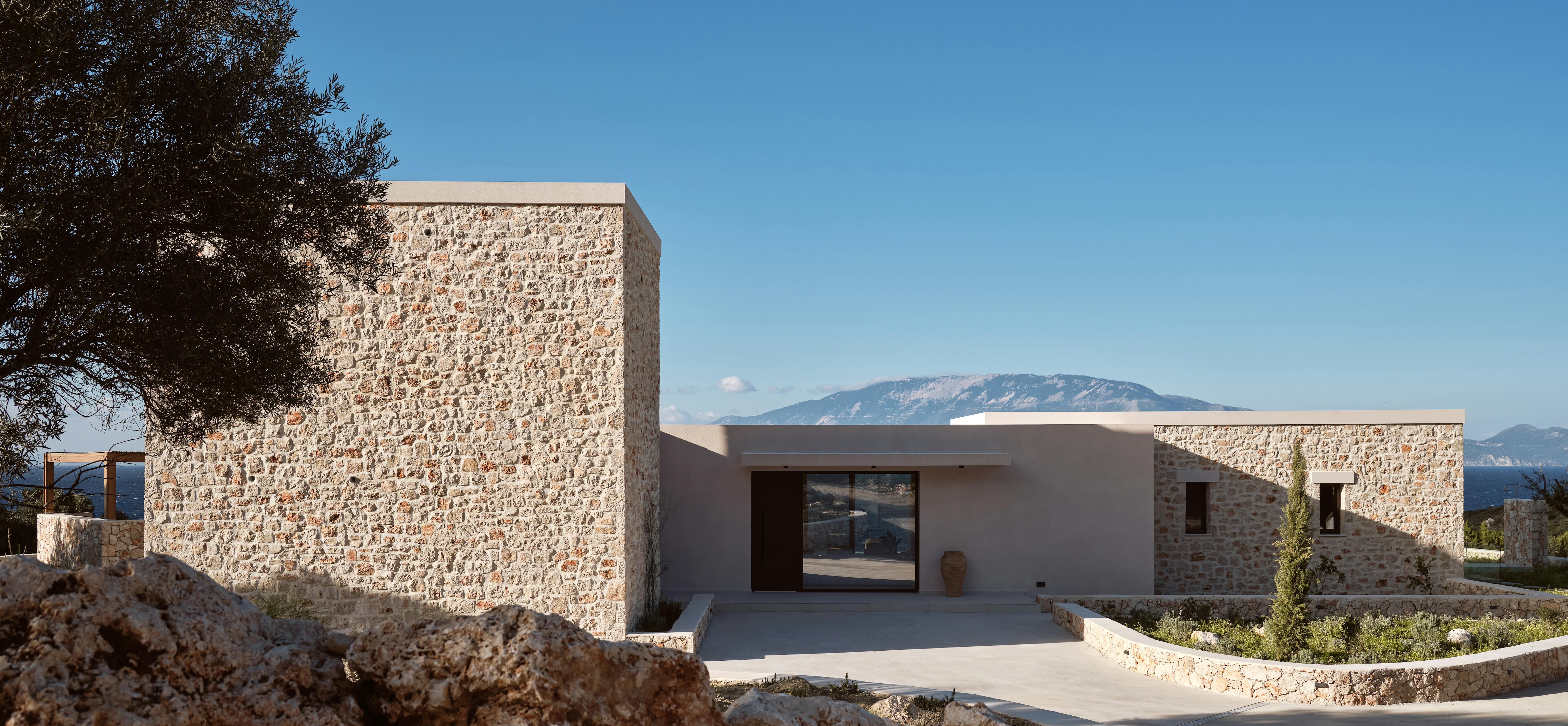 Stone-clad modern villa exterior with mixed materials of local stone and smooth plaster set into hillside with native Mediterranean vegetation.