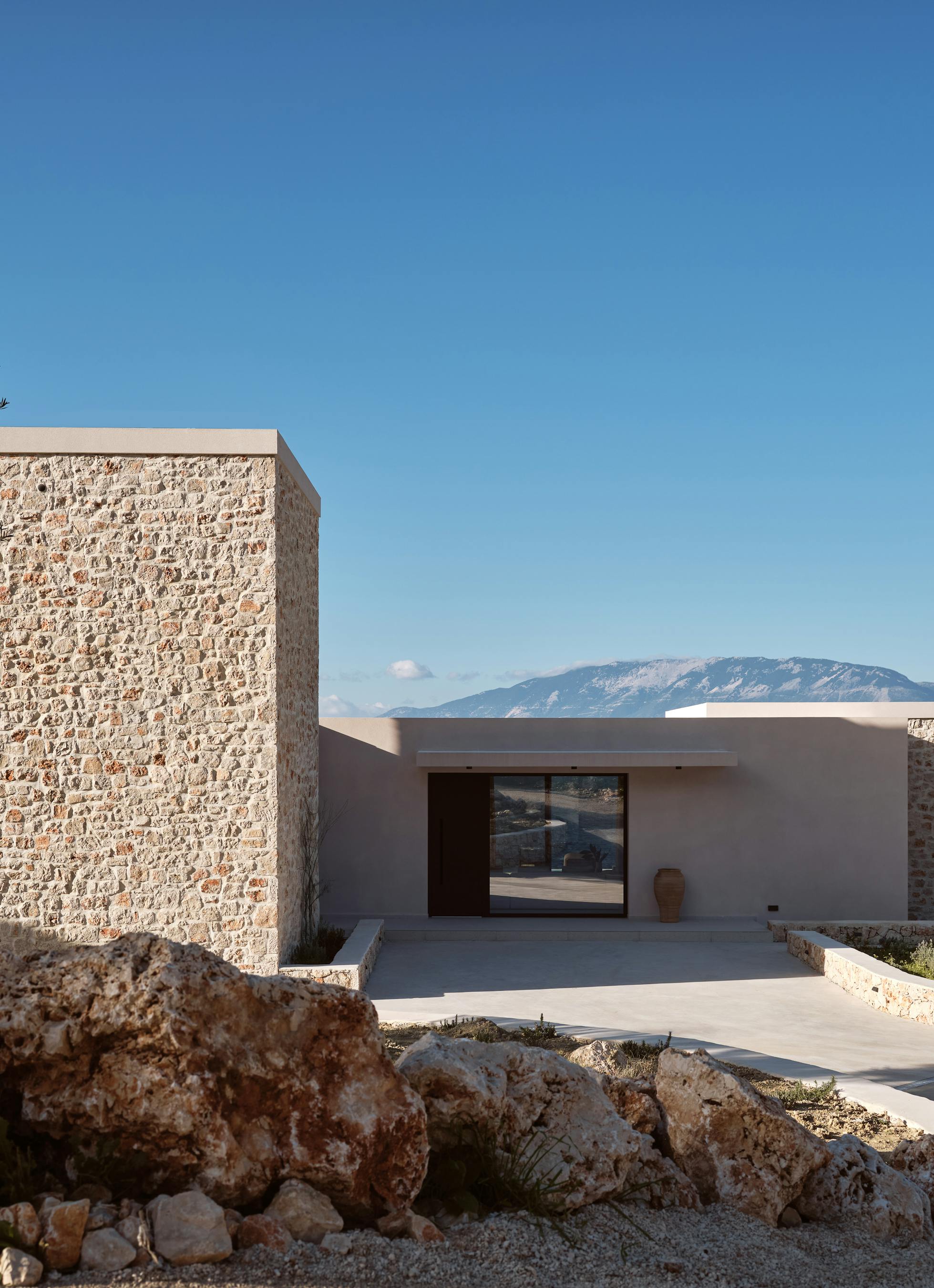 Stone-clad modern villa exterior with mixed materials of local stone and smooth plaster set into hillside with native Mediterranean vegetation.