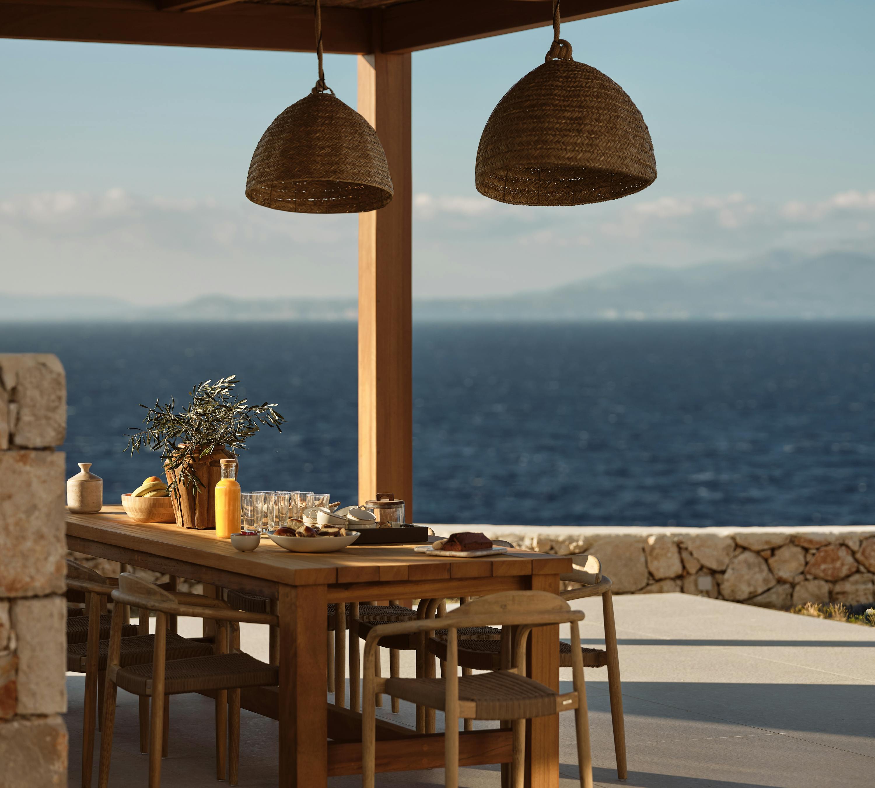 Outdoor dining area under wooden pergola with woven pendant lights, set table with breakfast items, and panoramic Mediterranean Sea views at golden hour.