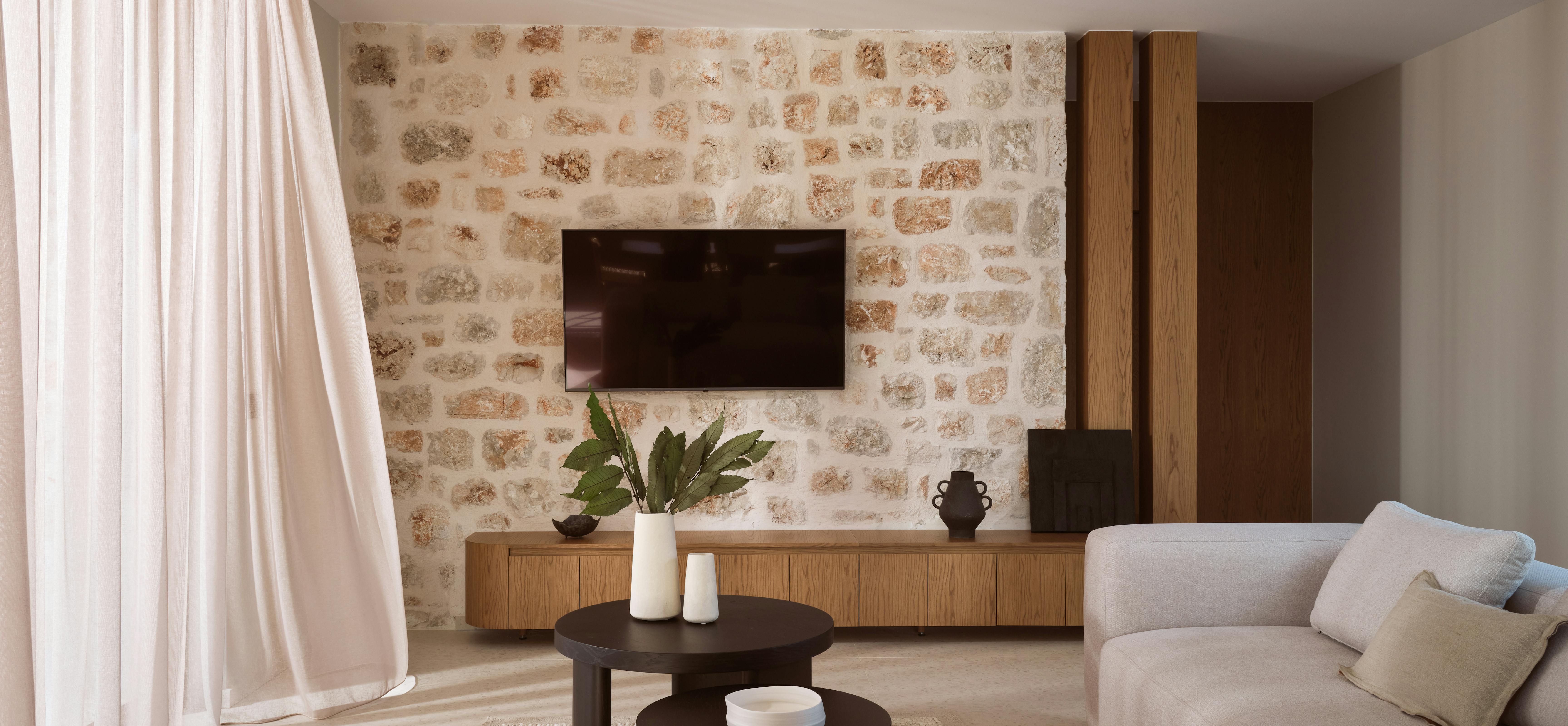 Contemporary living room featuring exposed stone accent wall, floating oak media console, modular sofas, and flowing white curtains with natural sunlight.