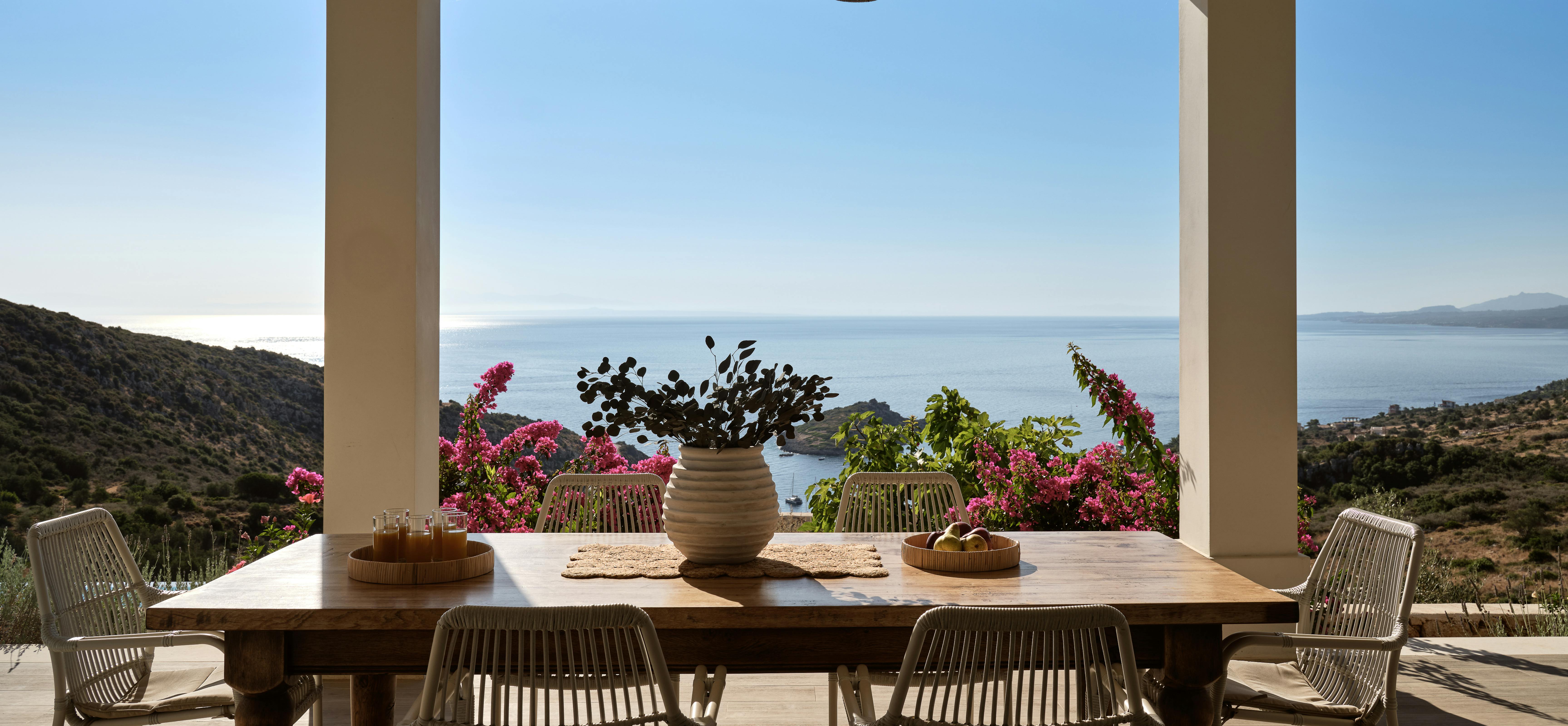 Open-air dining terrace with rustic wooden table, woven chairs, ceramic vessels with dried botanicals, and triple-arch openings framing Mediterranean Sea and coastal views.