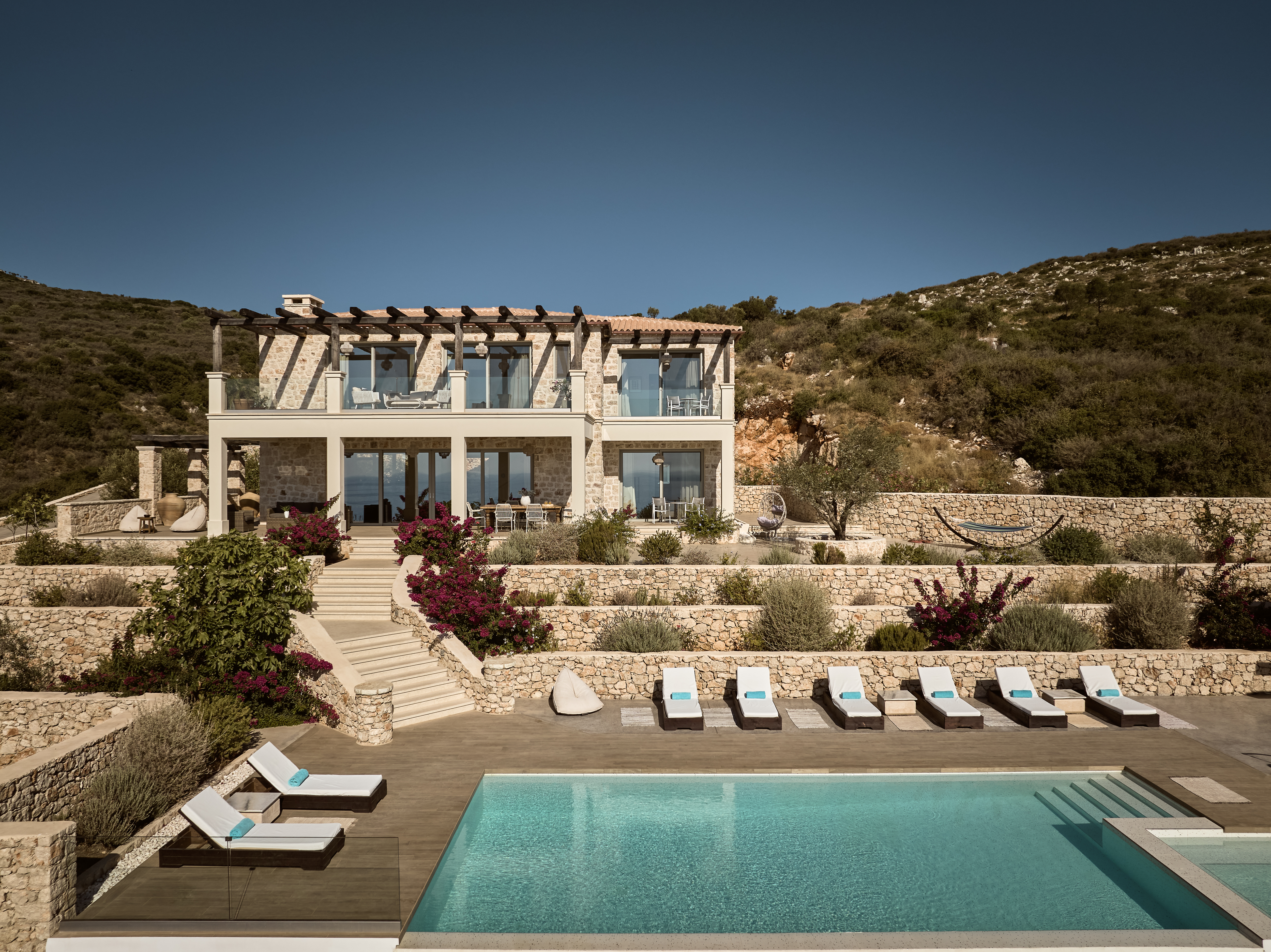 Two-story Mediterranean stone villa with wooden pergola rooftop terrace, terraced landscaping with bougainvillea, infinity pool, and sun loungers overlooking hillside.
