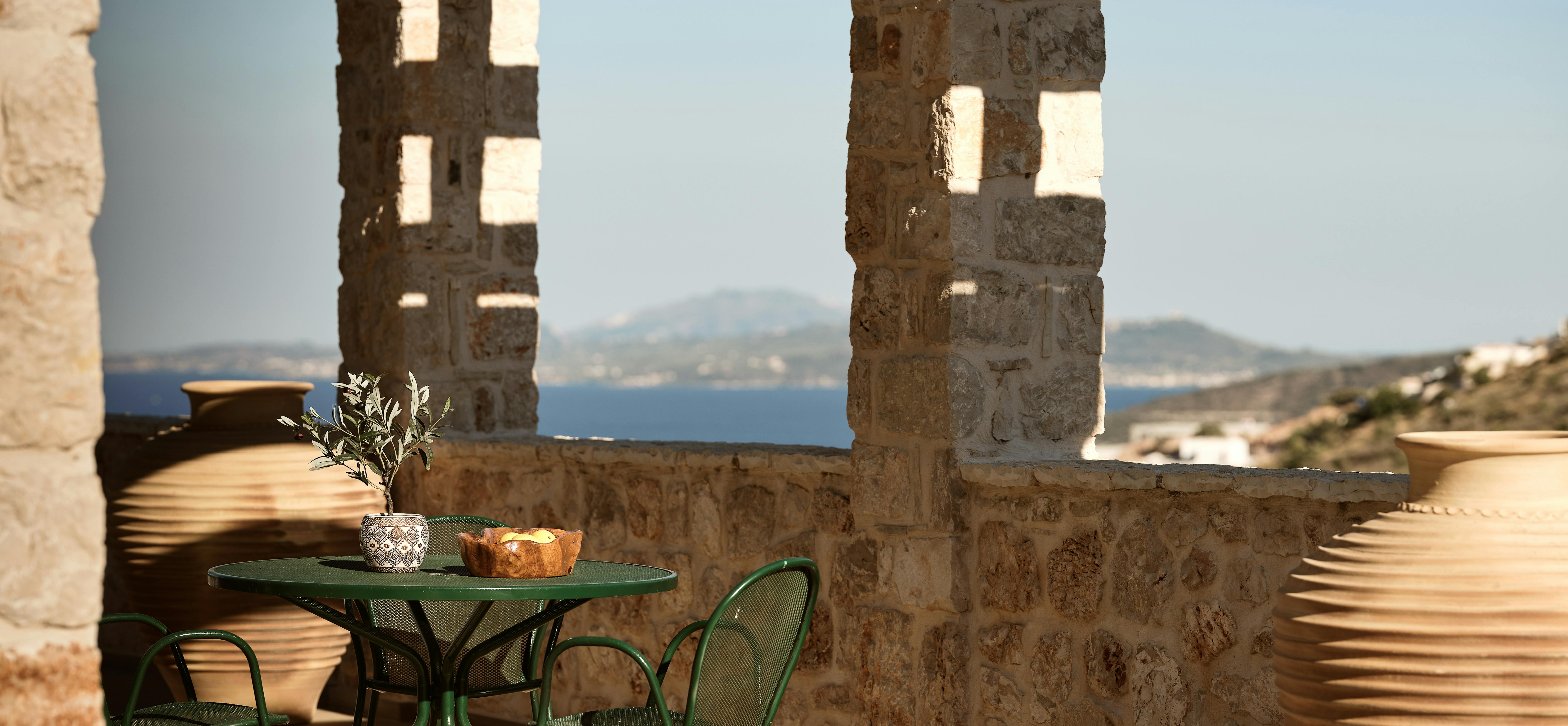 Intimate terrace bistro setting with green metal table and chairs under rustic stone columns framing distant island and sea views.