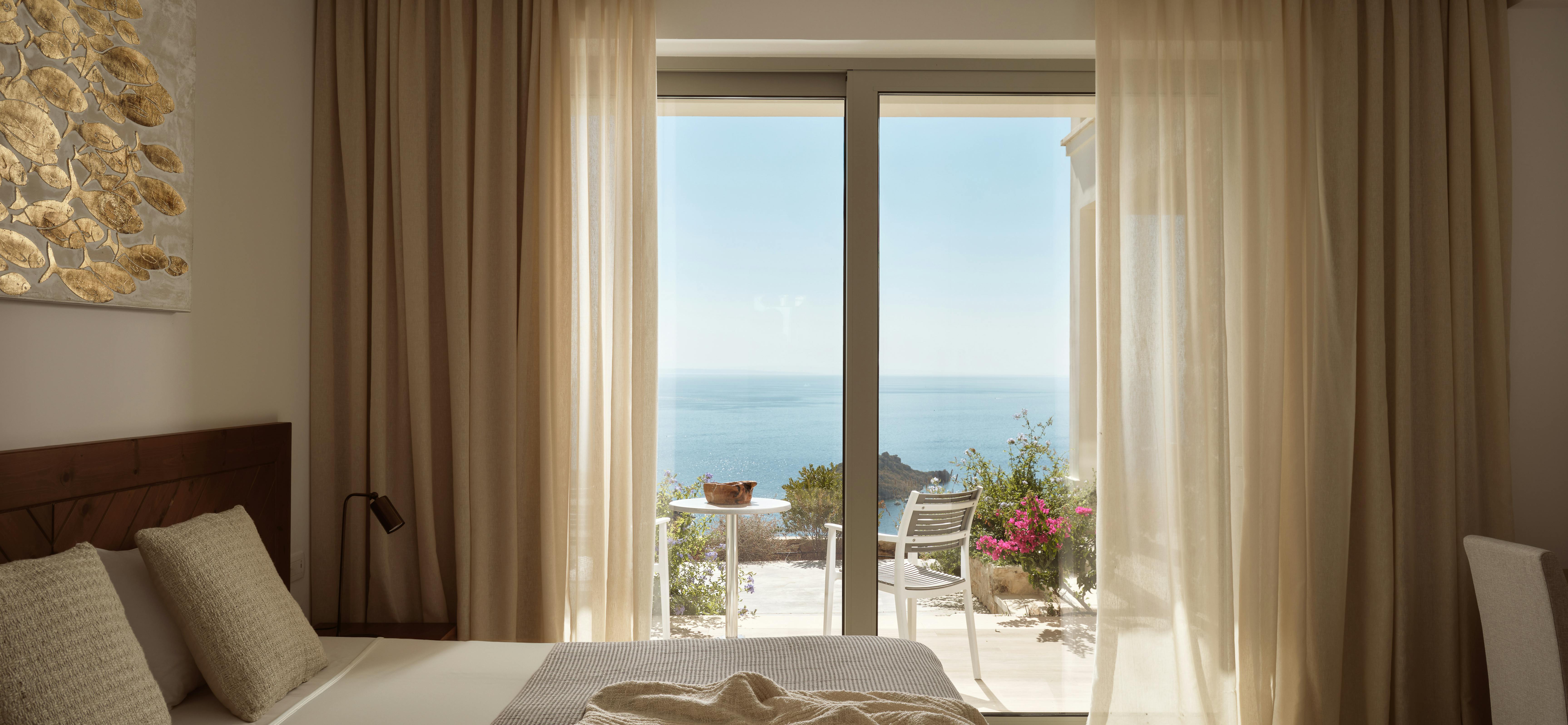Bright bedroom with golden fish artwork, wooden chevron headboard, layered neutral bedding, and sliding glass doors opening to sunny terrace with ocean views.