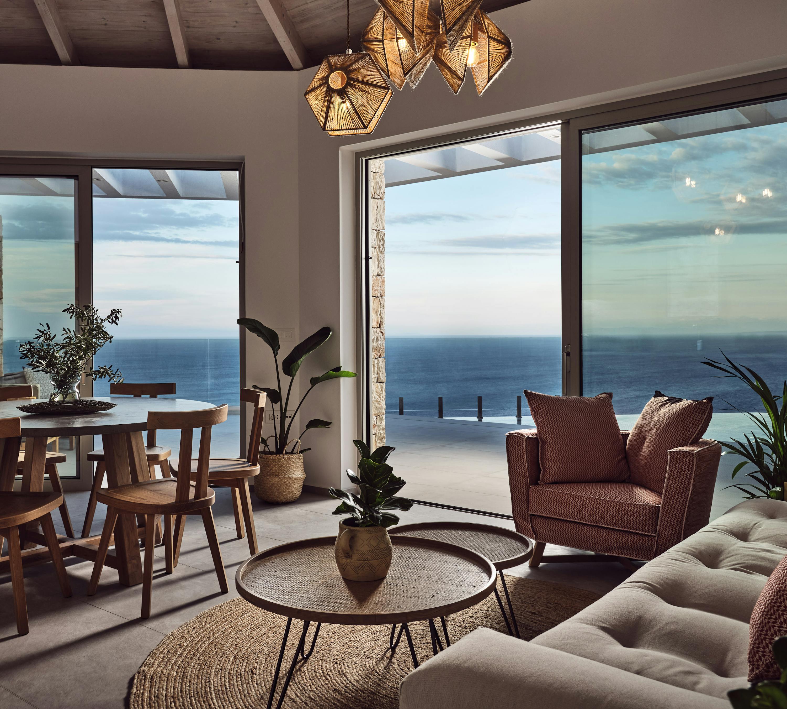 Open-plan living space with wooden dining set, terra-cotta textured armchair, tufted sofa, geometric pendant lights, and panoramic glass doors framing ocean and terrace views.