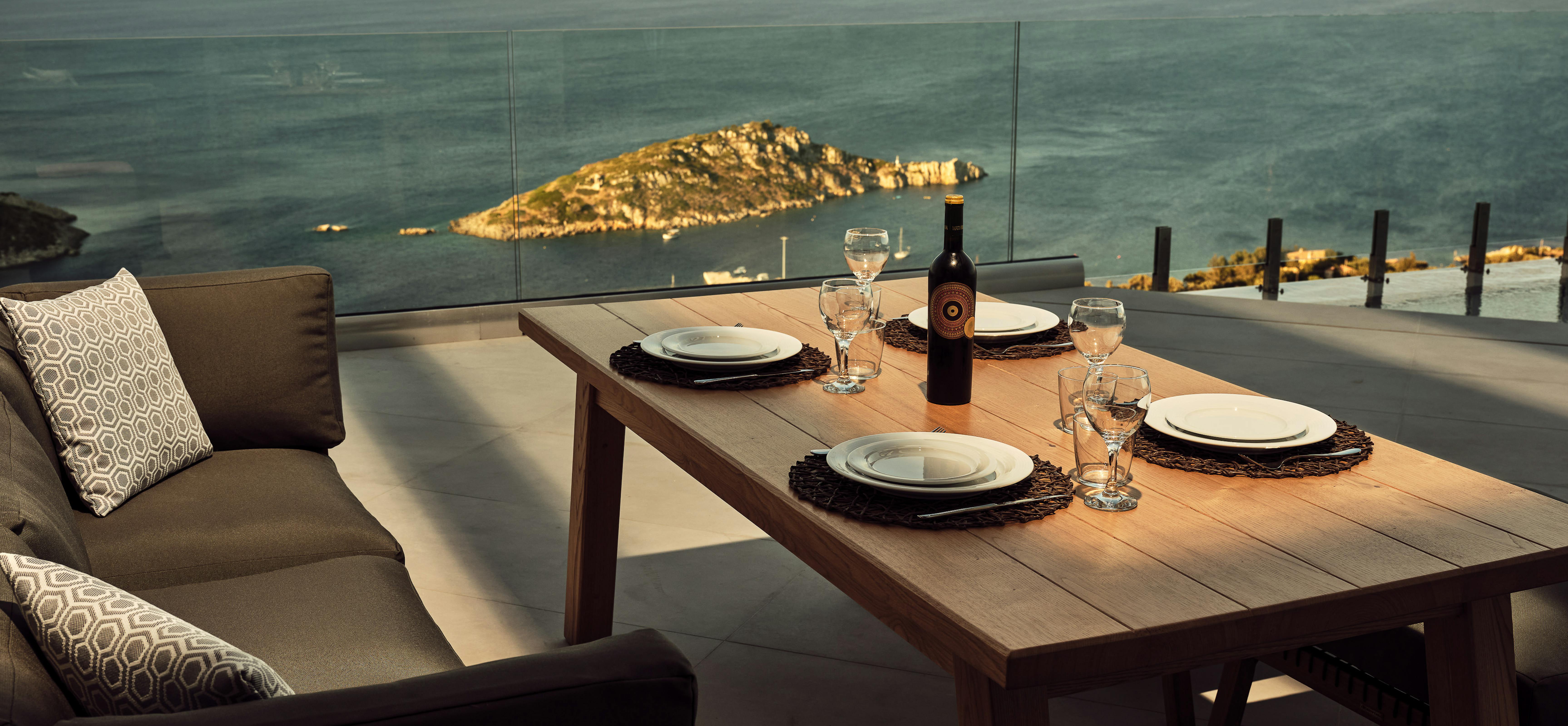 Golden hour terrace dining with wooden table set for three, woven placemats, wine bottle, cushioned bench seating, and glass railing framing rocky island and glowing sea.