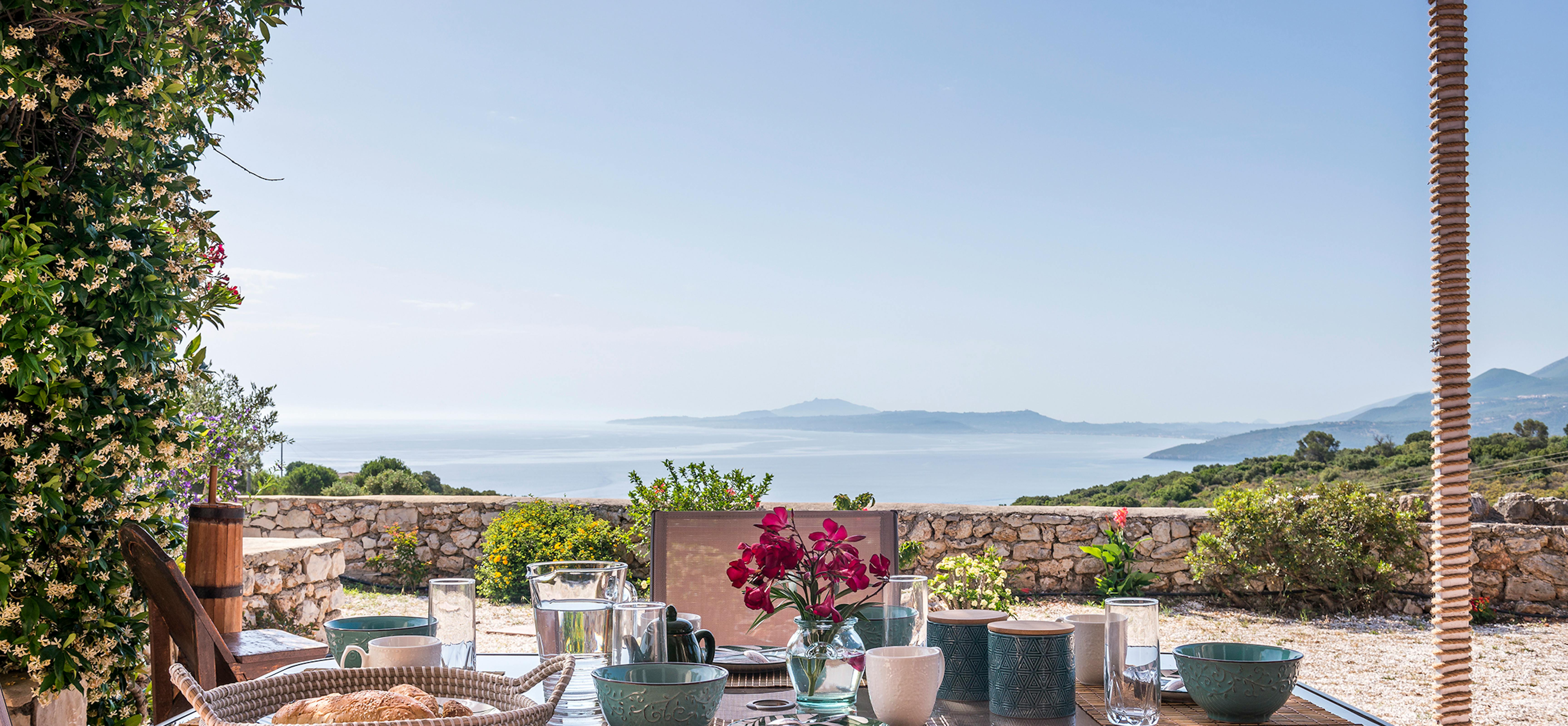 Outdoor breakfast table under reed pergola set with colourful ceramic bowls, glass water pitcher, fresh bougainvillea, and panoramic views of blue Mediterranean Sea and distant islands.