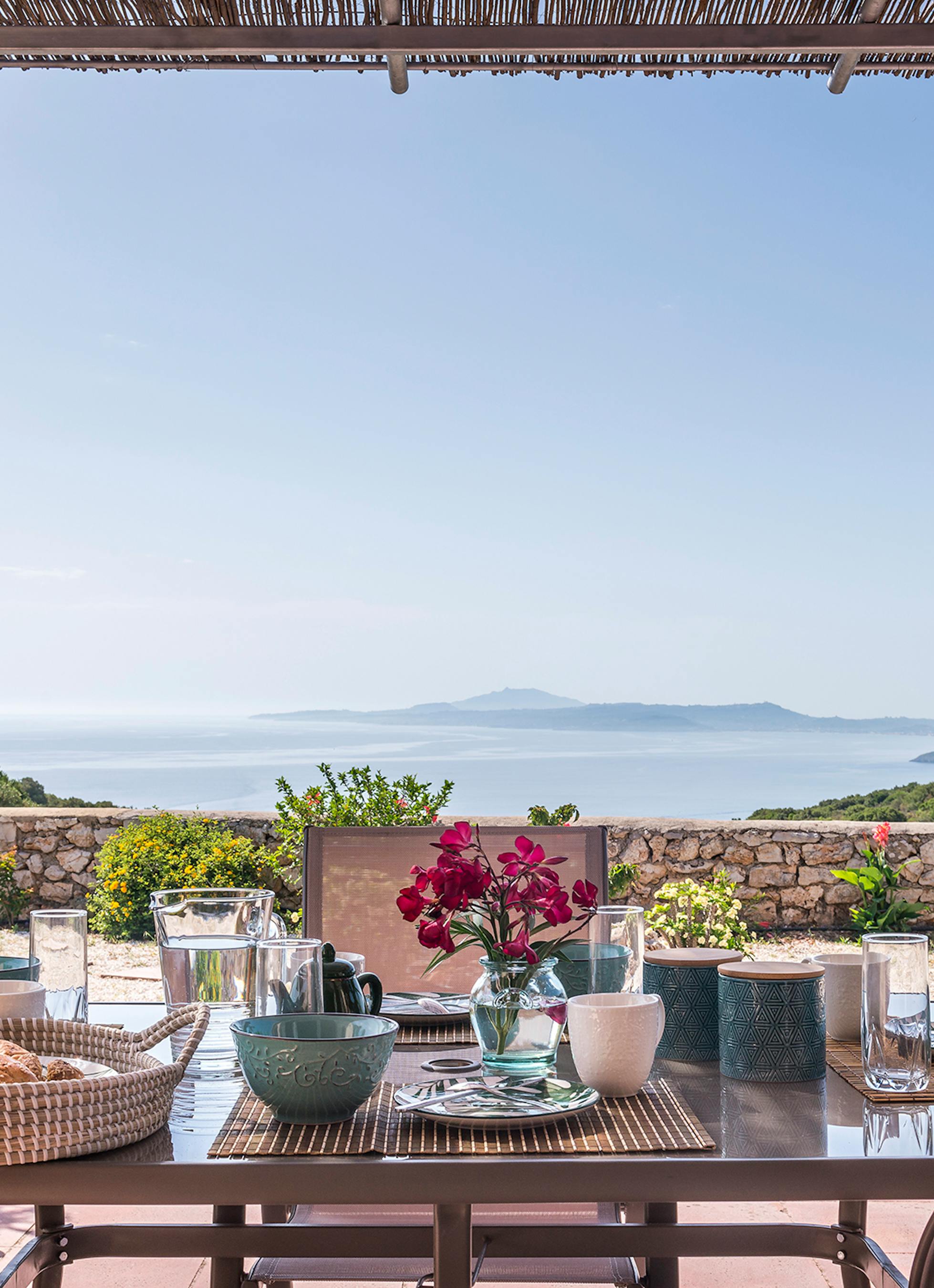 Outdoor breakfast table under reed pergola set with colourful ceramic bowls, glass water pitcher, fresh bougainvillea, and panoramic views of blue Mediterranean Sea and distant islands.