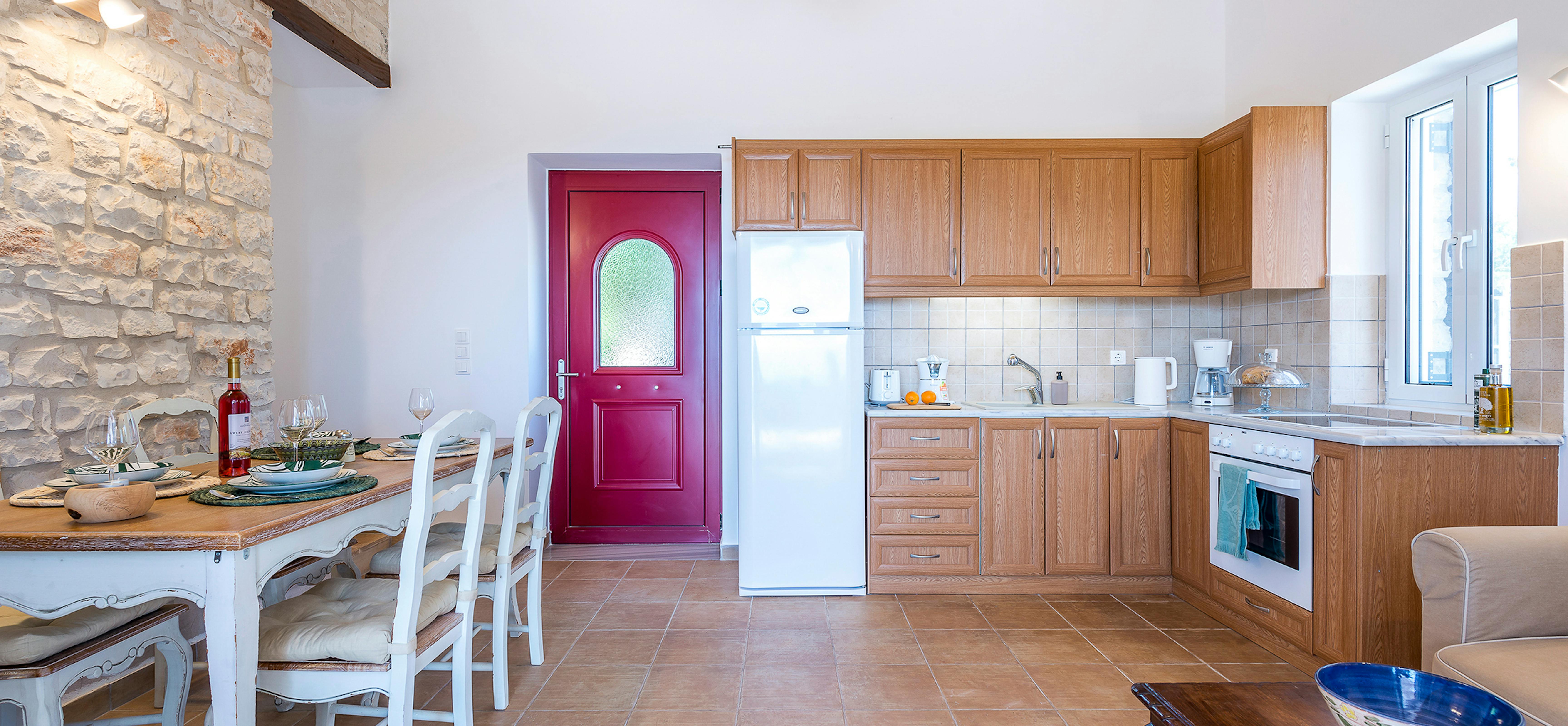 Cottage kitchen-dining room with vaulted wood ceiling, exposed stone wall, oak cabinetry, vibrant magenta entry door with arched glass, white dining set, and terracotta tile flooring.