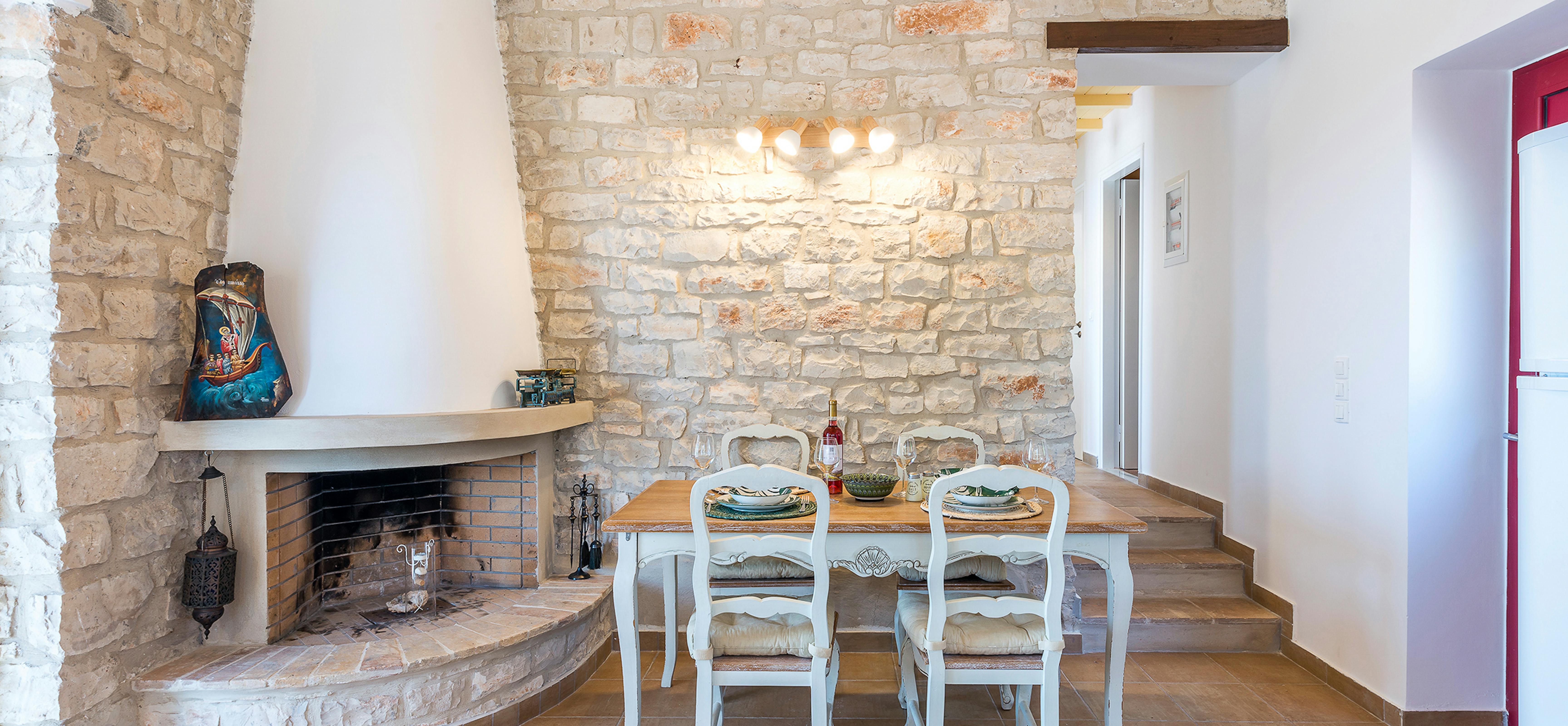 Cottage dining area with dramatic stone accent wall, rustic fireplace hearth, white French Provincial dining chairs, and decorative blue ceramic ship sculpture on wooden shelf.
