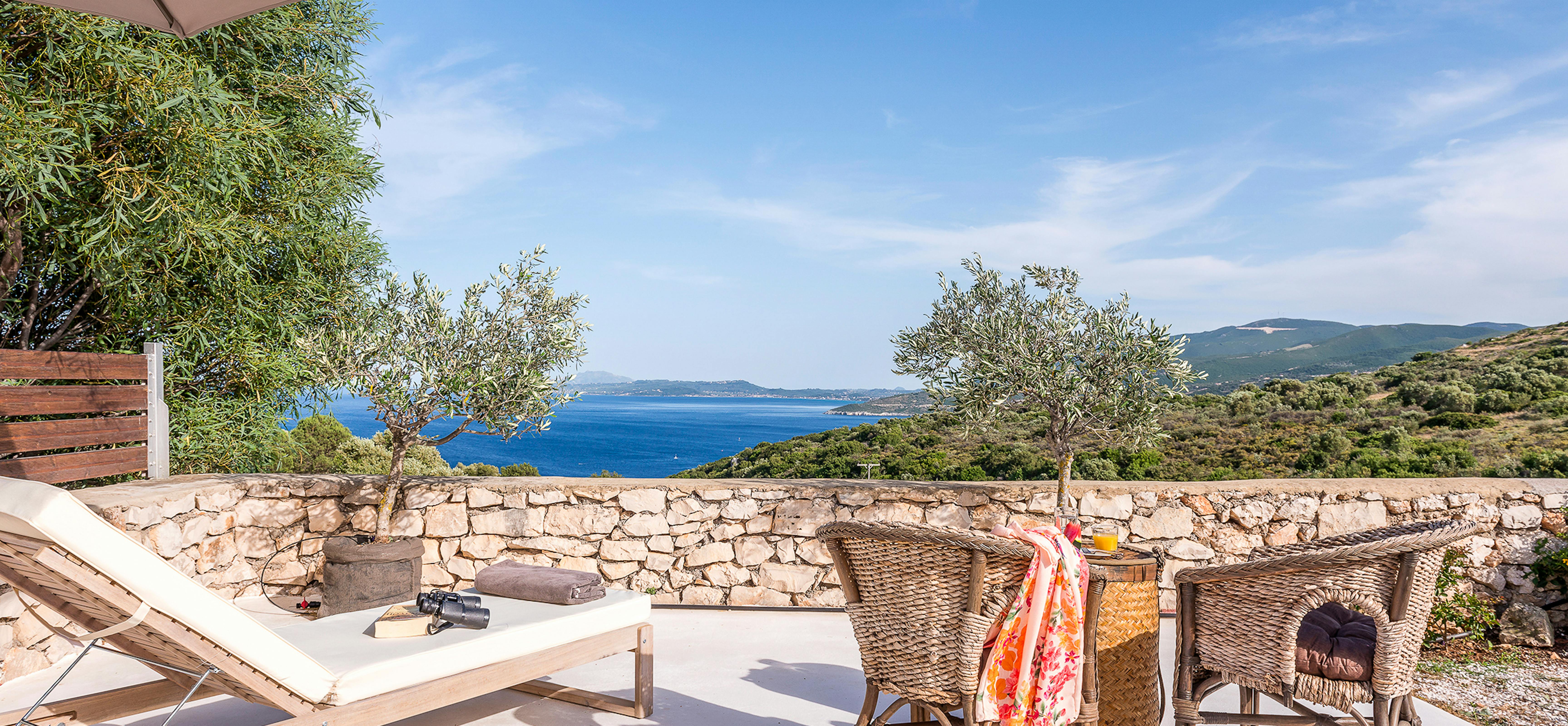 Sun-drenched terrace with white lounger, wicker dining set draped with colourful towel, taupe umbrella, stone retaining walls, olive trees, and sweeping coastal panorama.