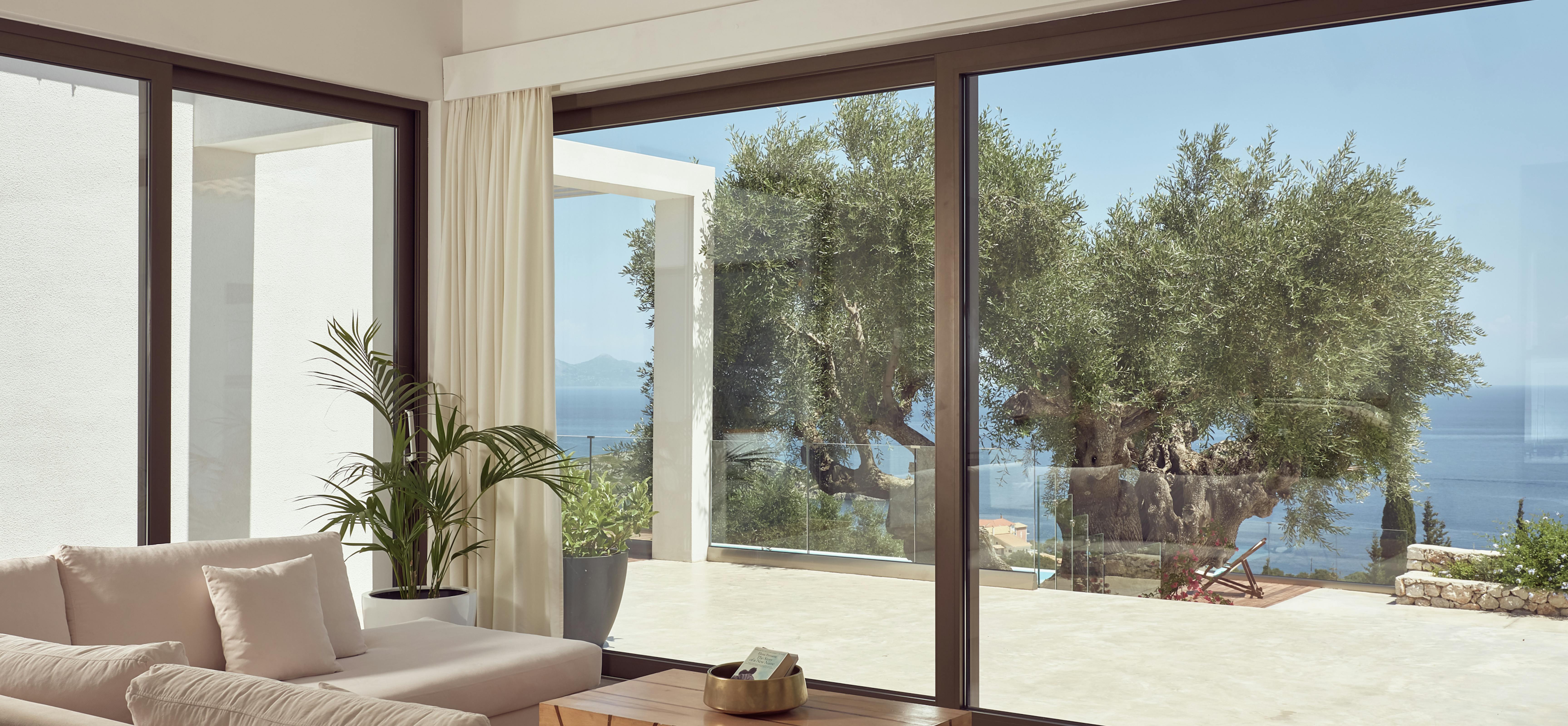 Modern living room with floor-to-ceiling windows overlooking a Mediterranean terrace with ancient olive tree and sea views.