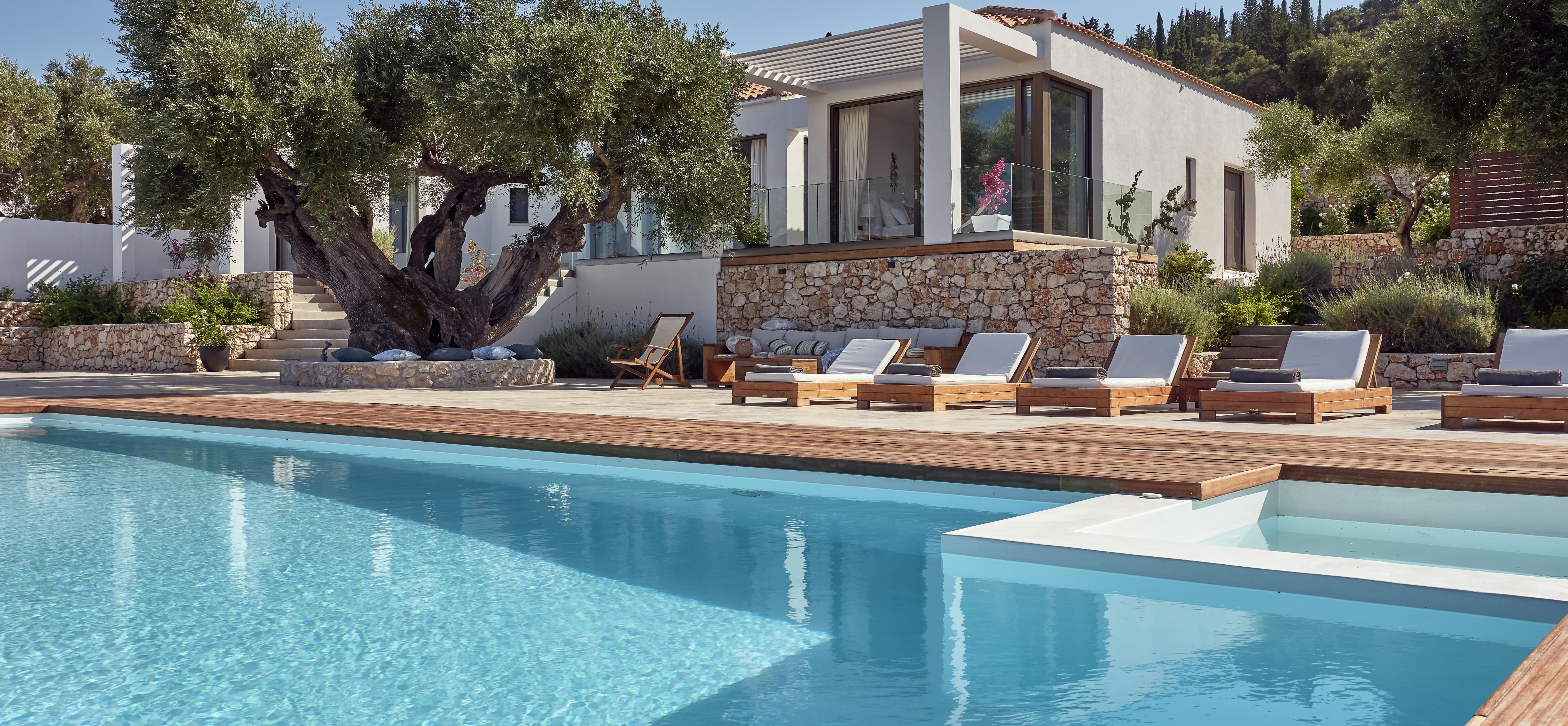 Luxury villa exterior with infinity pool, wooden deck, sun loungers, and ancient olive trees at dusk.