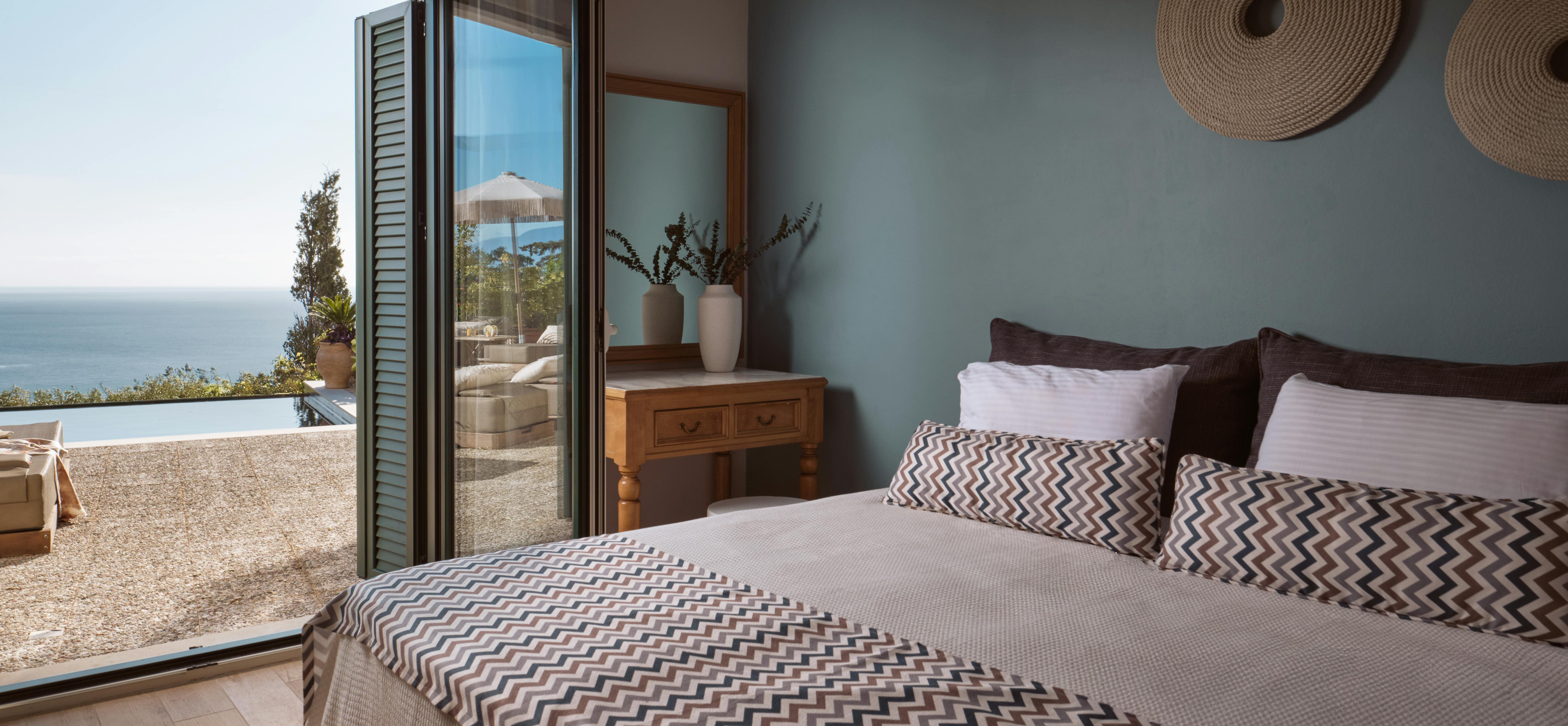 A serene bedroom with soft blue wall features chevron-patterned bedding and woven wall art. Large windows and doors open to a poolside terrace with views of outdoor furniture and umbrella.
