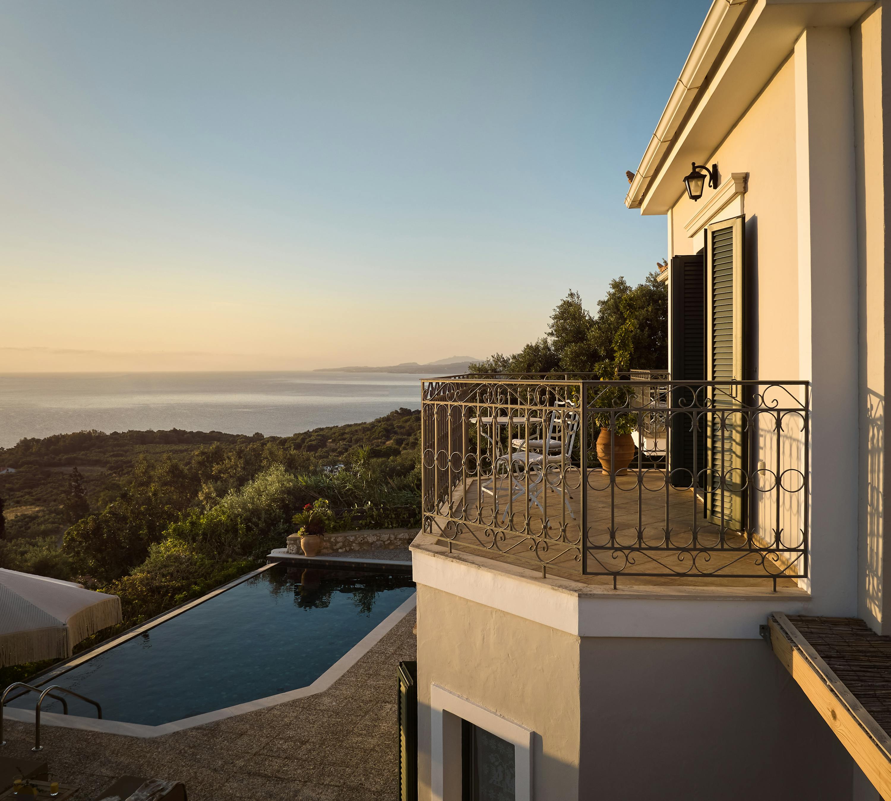 A villa balcony captures golden hour light with wrought iron railings overlooking rolling hills and distant sea. The terracotta-roofed building exemplifies classic Mediterranean architecture.
