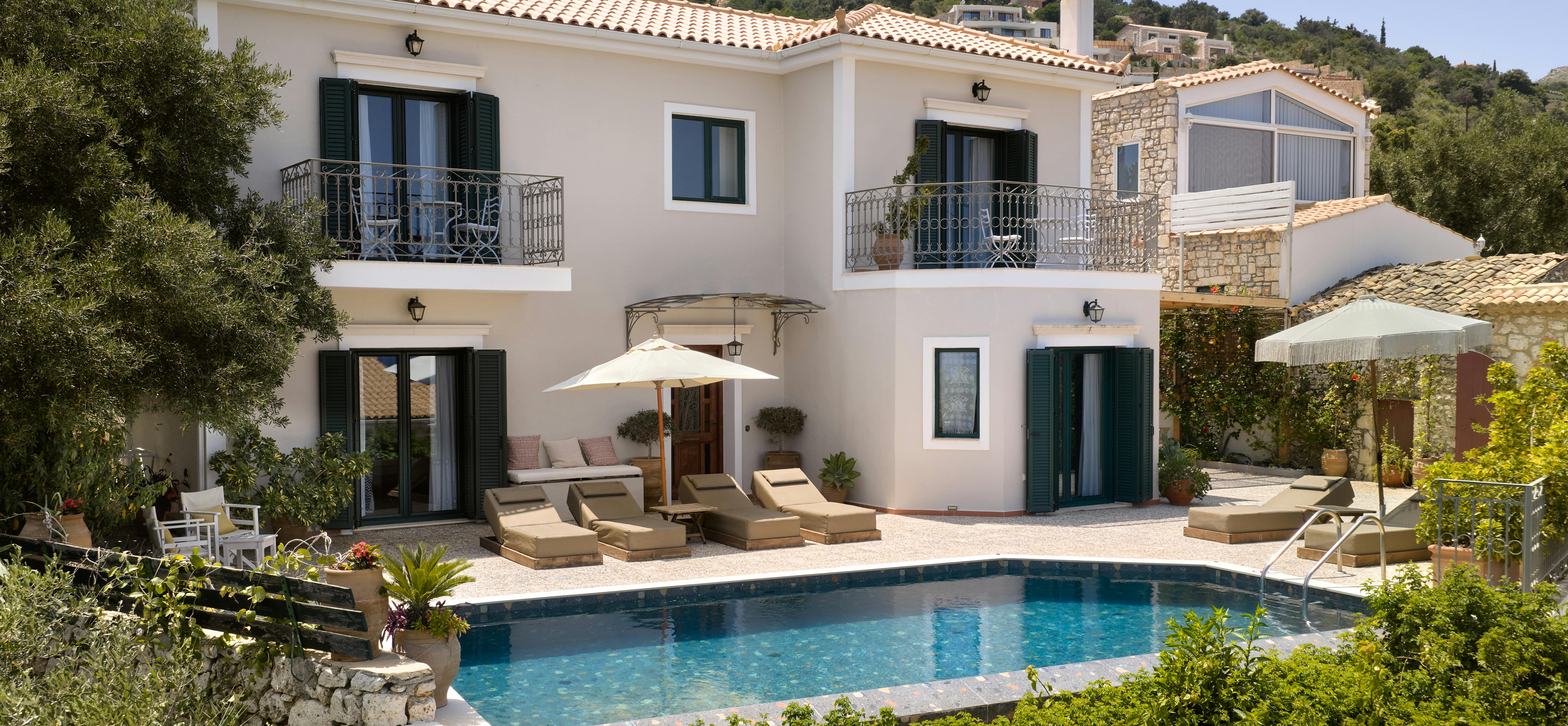 A Mediterranean villa with terracotta roof tiles sits on a hillside overlooking a rectangular swimming pool. Wooden lounge furniture and umbrellas are arranged on the stone terrace surrounded by lush landscaping.