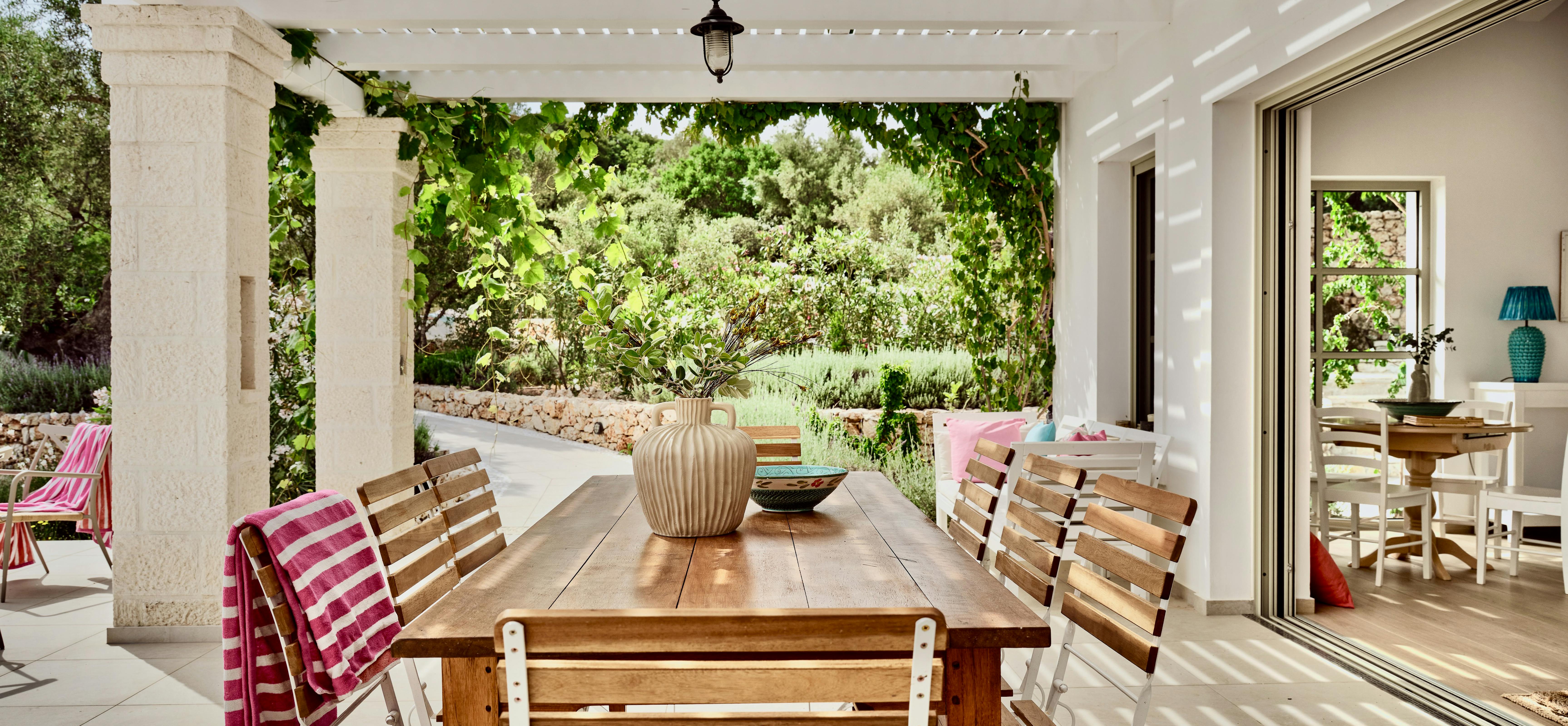 Covered outdoor dining terrace with stone columns and white pergola ceiling. A wooden table with folding chairs overlooks gardens and trees.