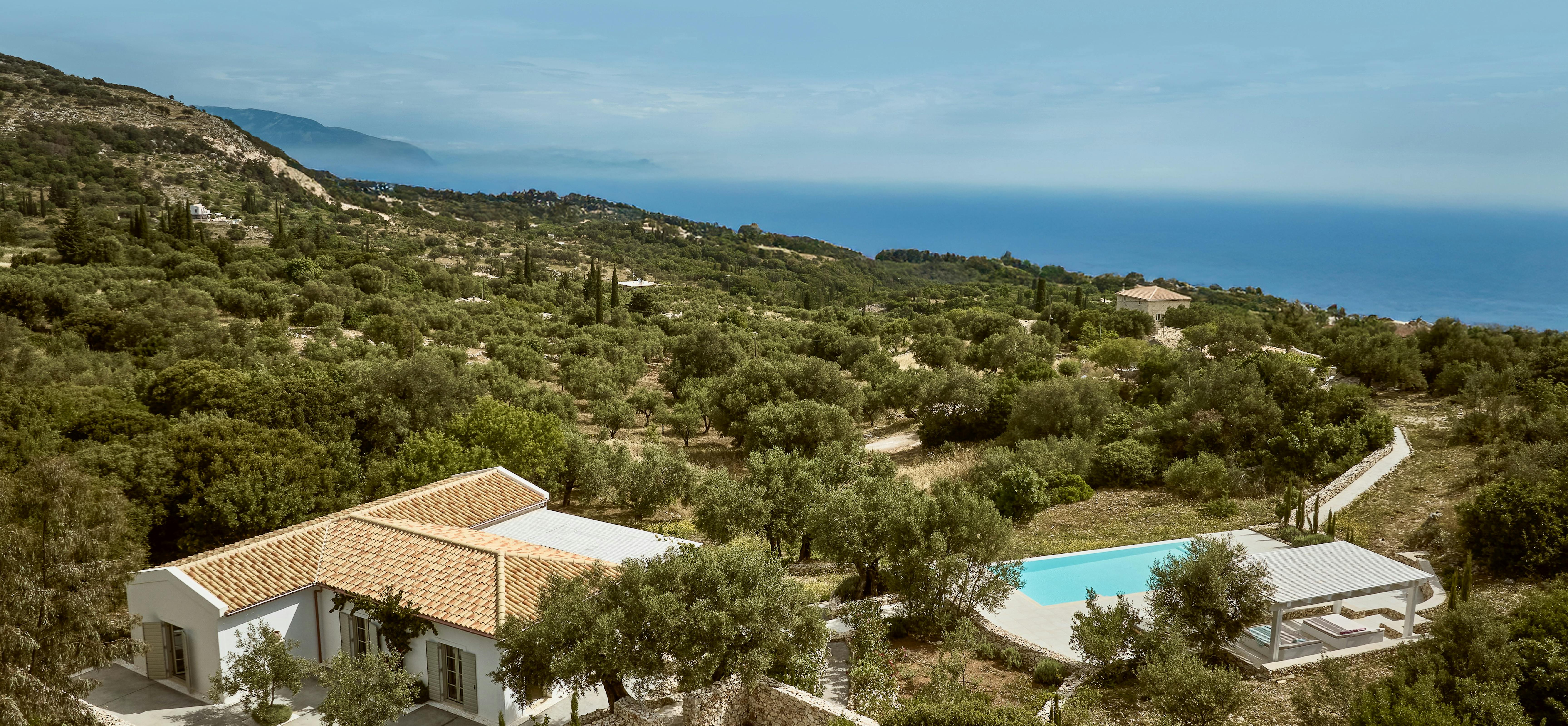 Aerial view of Mediterranean villa with terracotta roof nestled in hillside overlooking the sea. Swimming pool and white pergola structures complement the natural landscape of olive trees and cypress.