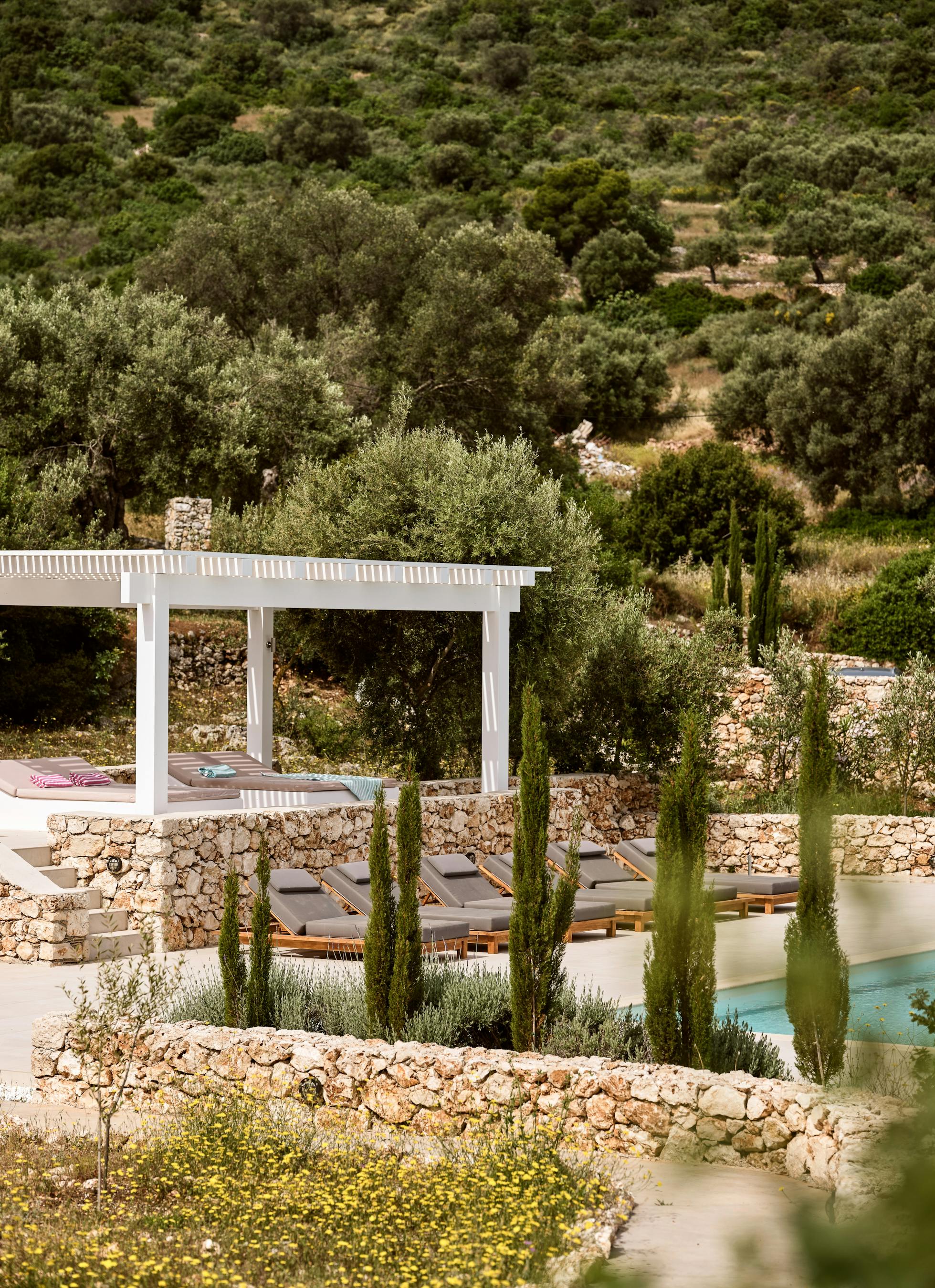 Pool area from elevated perspective showing integration with natural hillside terrain. Stone retaining walls and Mediterranean plantings blend architecture with landscape.