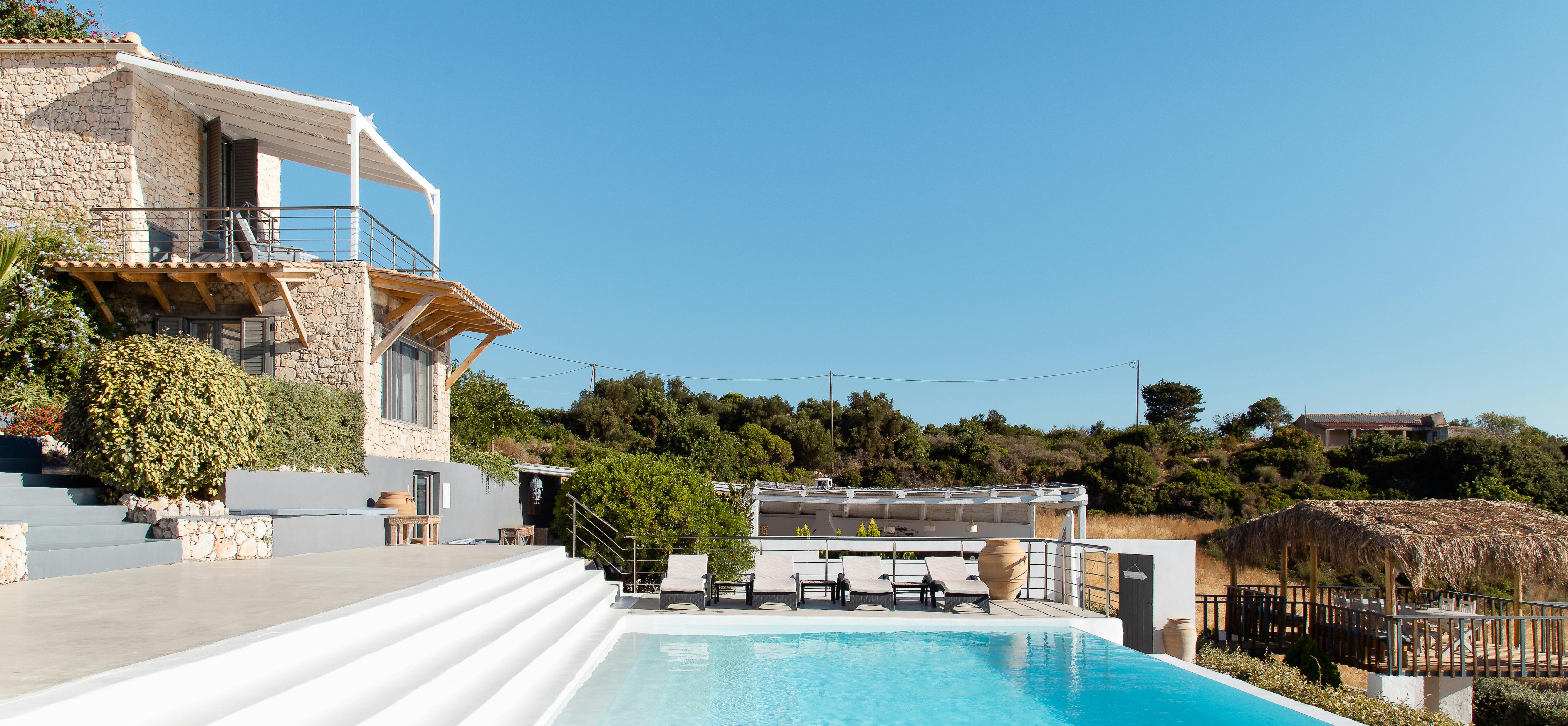 A large pool creates dramatic reflections against the turquoise Mediterranean Sea below. Modern white architecture contrasts with natural stone landscaping and coastal vegetation.