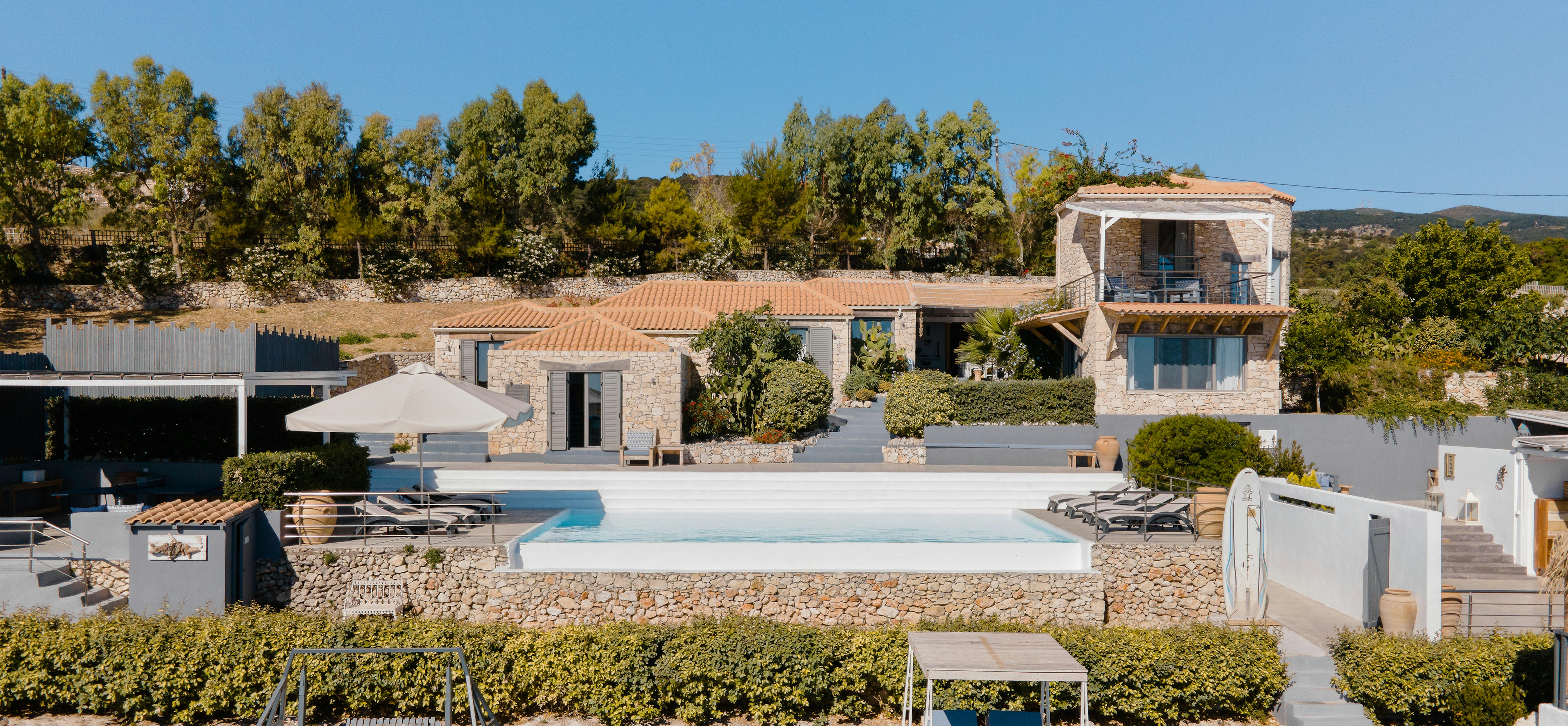 A Mediterranean hillside estate features multiple stone buildings with terracotta roofs surrounding a large rectangular swimming pool. The property includes terraced levels with an outdoor fitness area complete with exercise equipment and daybed.