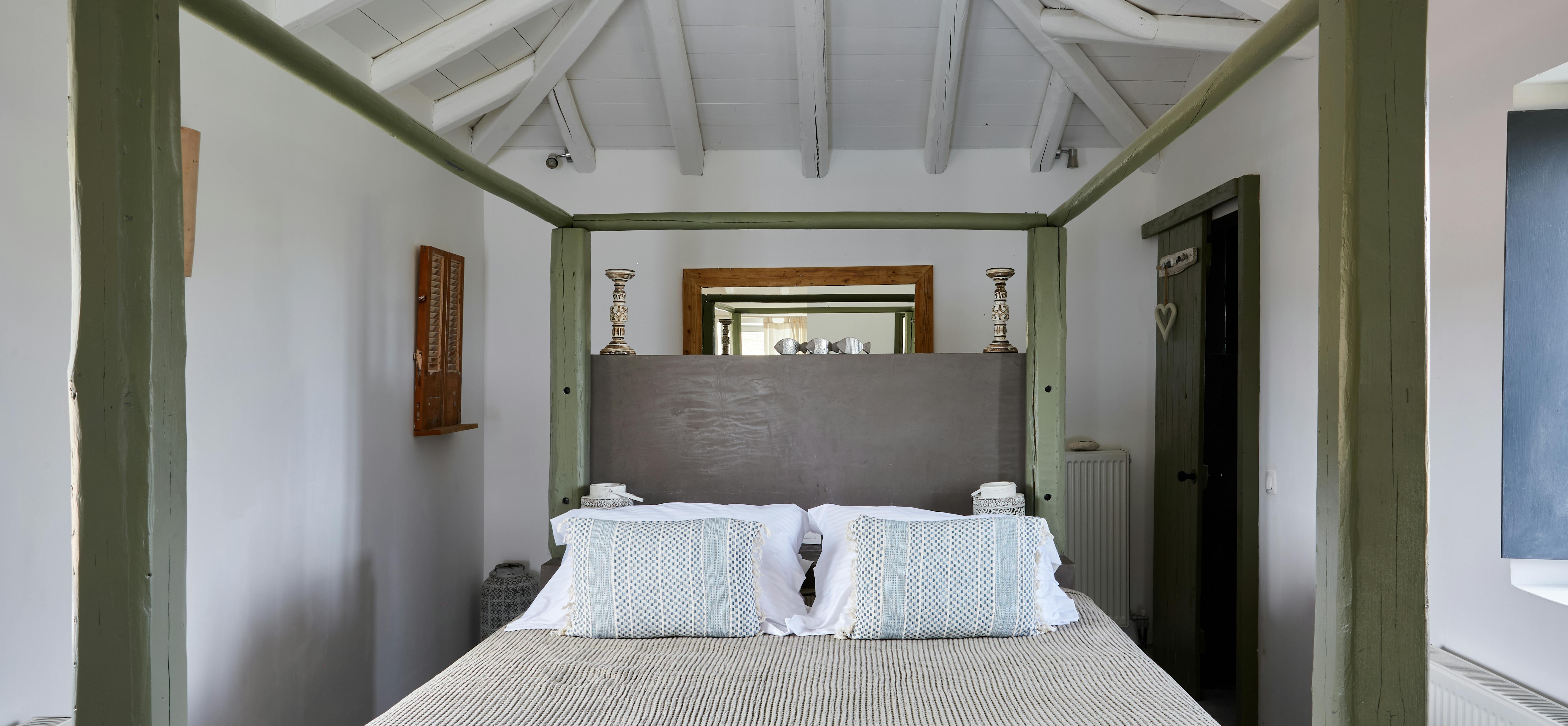 A rustic bedroom showcases a green four-poster bed frame with white linens and exposed ceiling beams. Traditional wooden shutters and ceramic accessories complement the Mediterranean-inspired interior design.