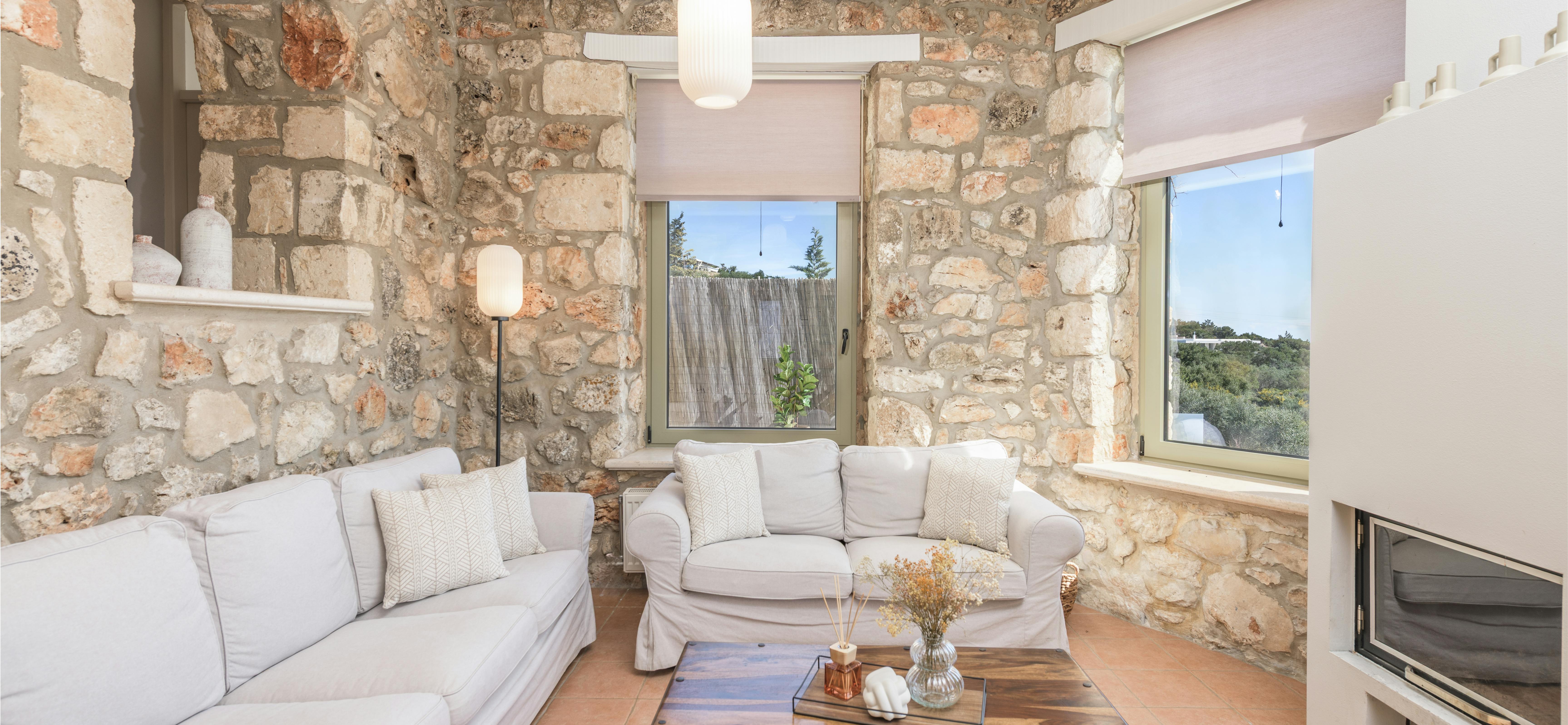 Â A living room features white slipcovered sofas arranged around a dark wooden coffee table beneath exposed ceiling beams. The neutral palette and natural textures create a relaxed atmosphere within the stone-walled space.