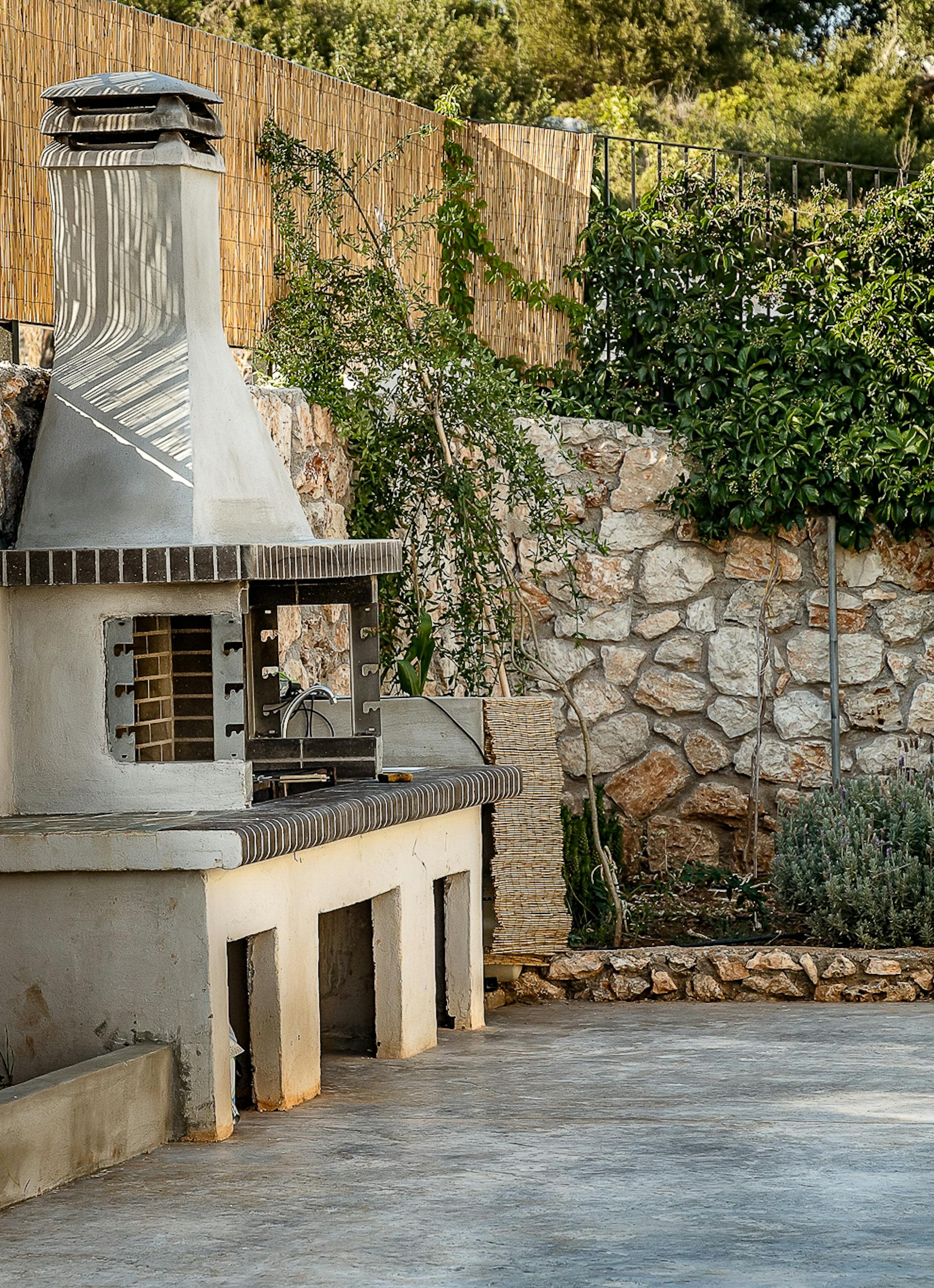 An outdoor kitchen area includes a built-in barbecue grill surrounded by stone walls and Mediterranean vegetation. The functional cooking space integrates seamlessly with the natural landscape and traditional stonework.