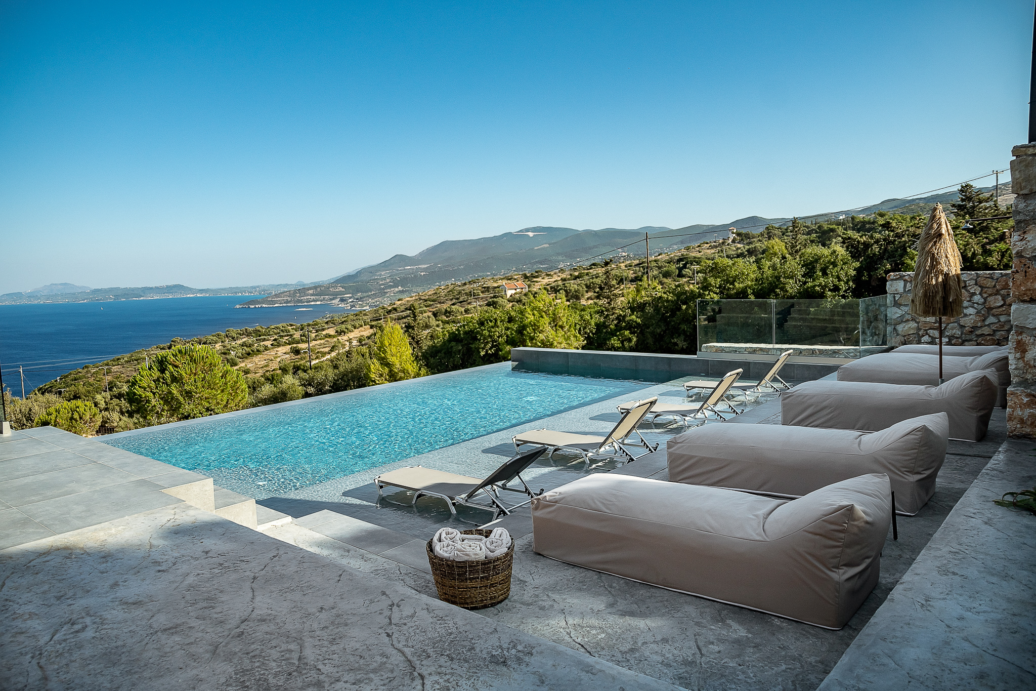 A hillside infinity pool terrace features large neutral cushions and lounge chairs positioned to maximise stunning Mediterranean sea views. The modern outdoor living space seamlessly blends luxury comfort with dramatic coastal scenery.