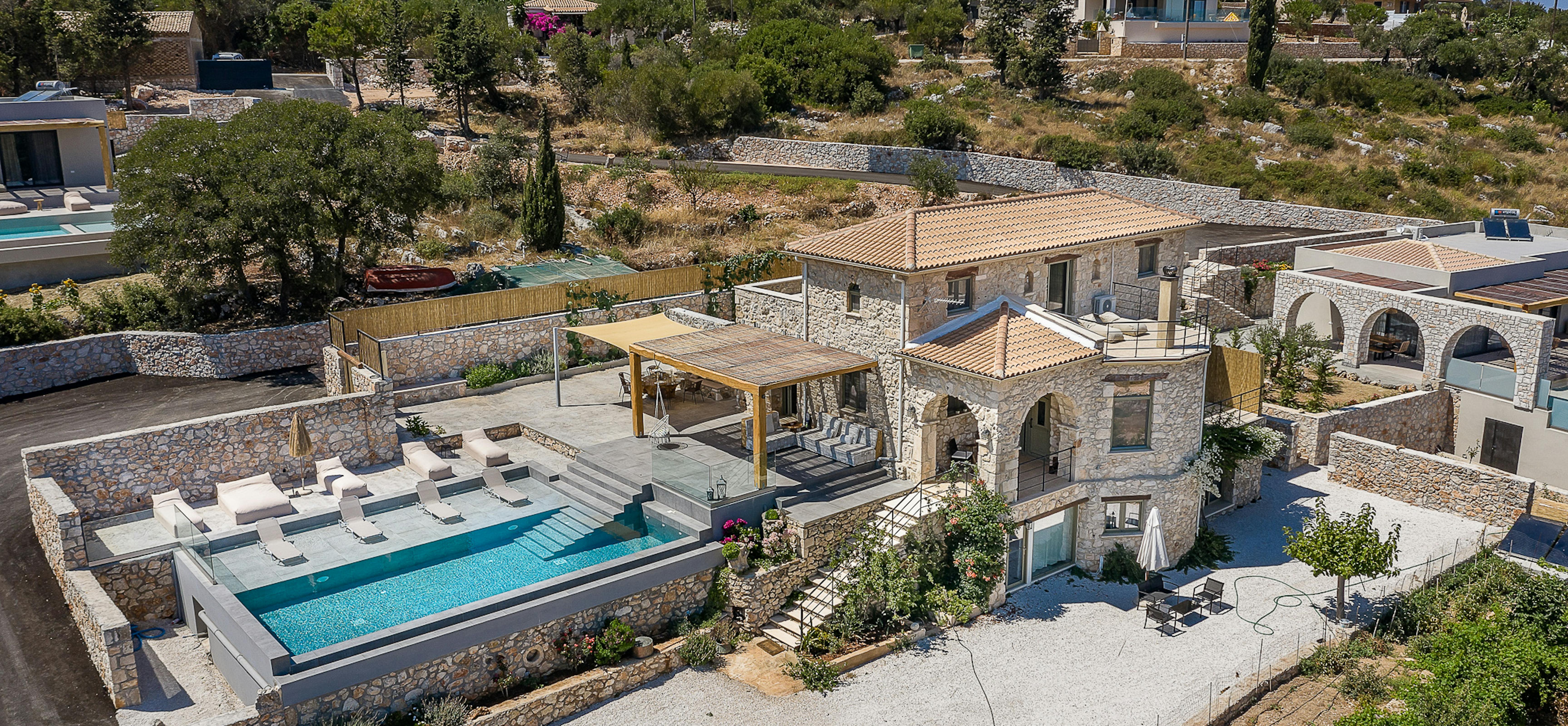 An aerial view reveals a stone villa complex with terracotta roofing, swimming pool, and multiple outdoor living areas. The Mediterranean estate integrates traditional architecture with modern amenities across terraced hillside grounds.