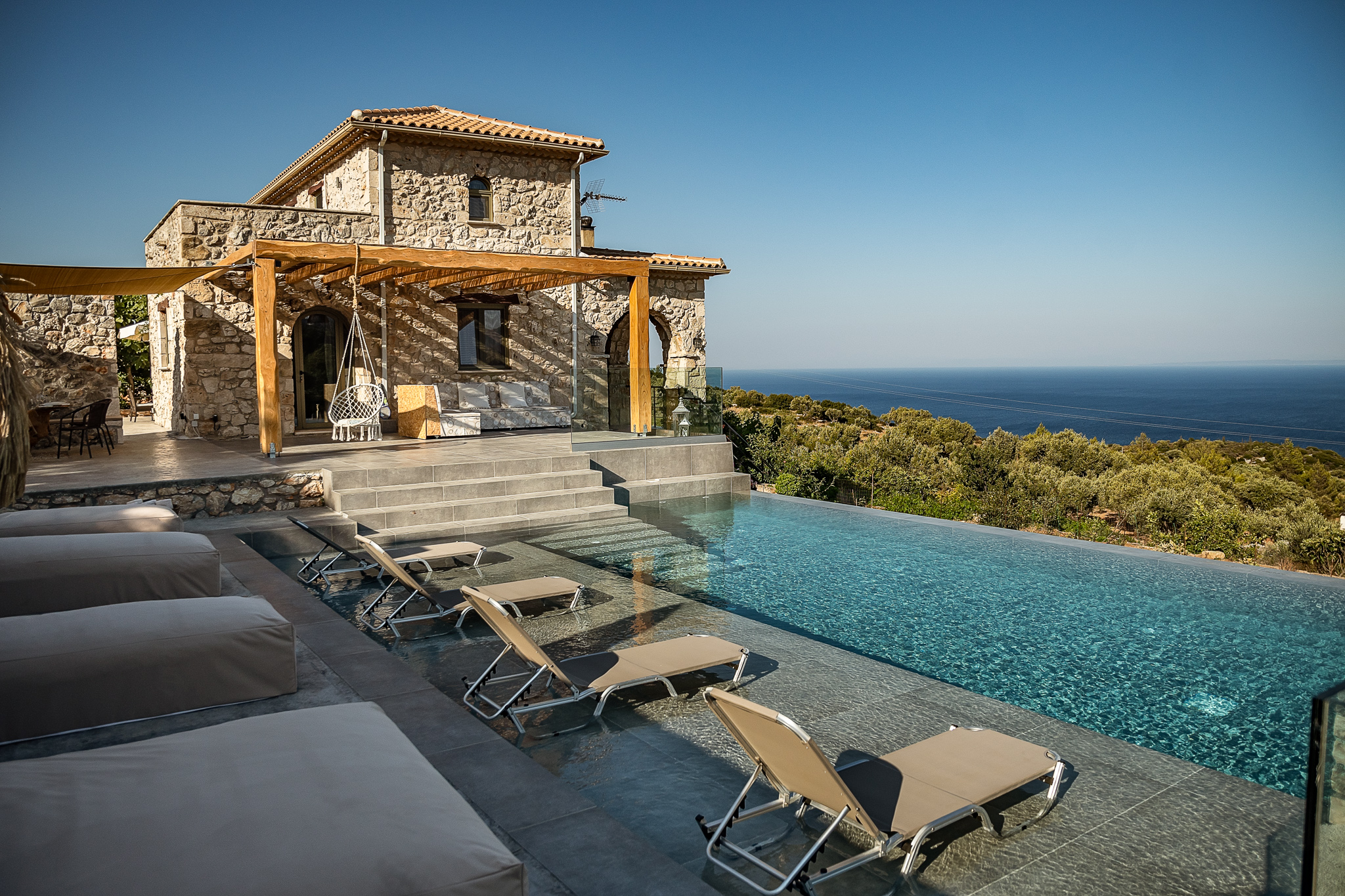 A stone Mediterranean villa overlooks an infinity pool with lounge chairs positioned on the deck for sea views. The traditional architecture features terracotta roofing and natural stone construction integrated into the coastal landscape.