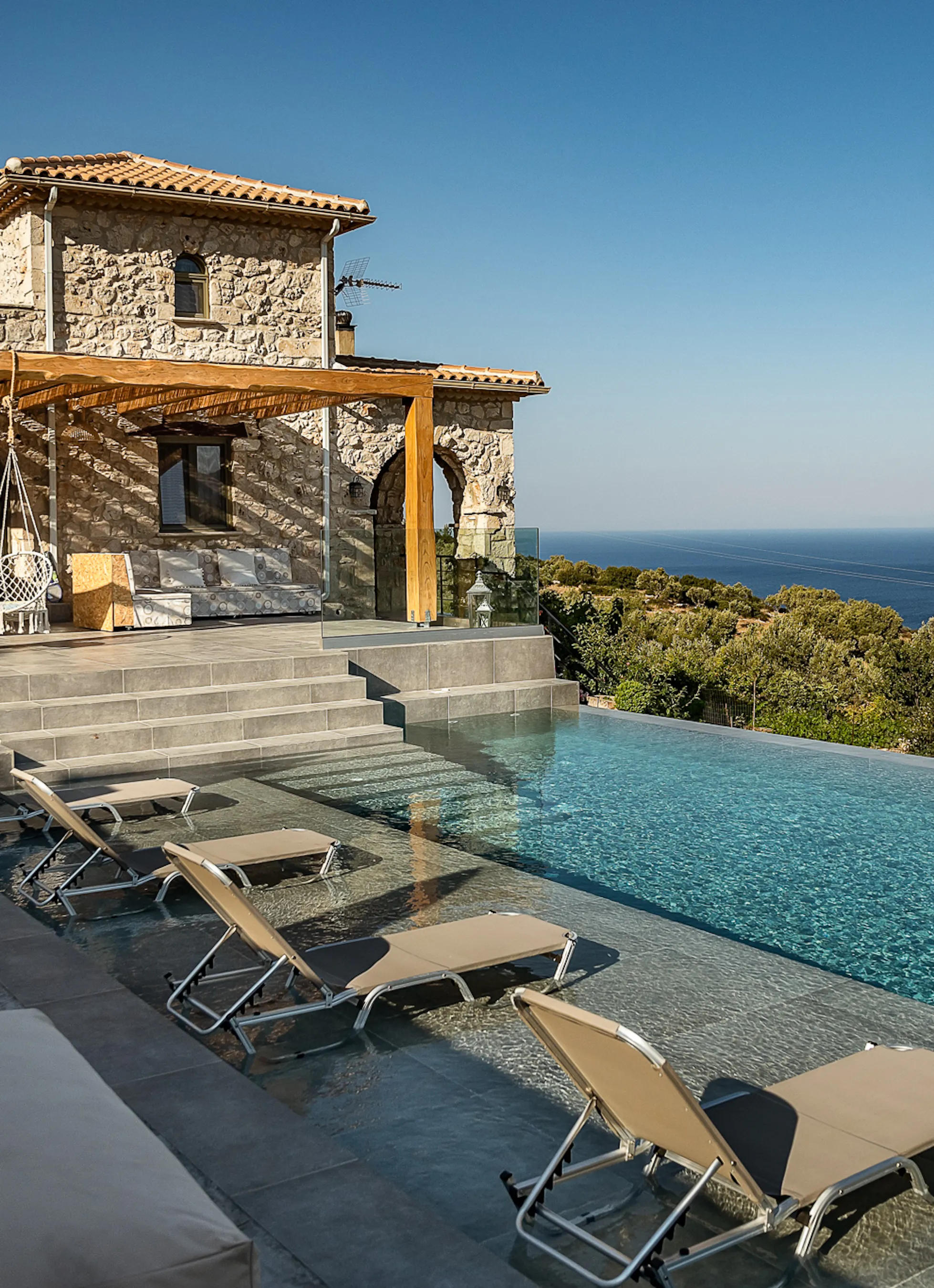 A stone Mediterranean villa overlooks an infinity pool with lounge chairs positioned on the deck for sea views. The traditional architecture features terracotta roofing and natural stone construction integrated into the coastal landscape.
