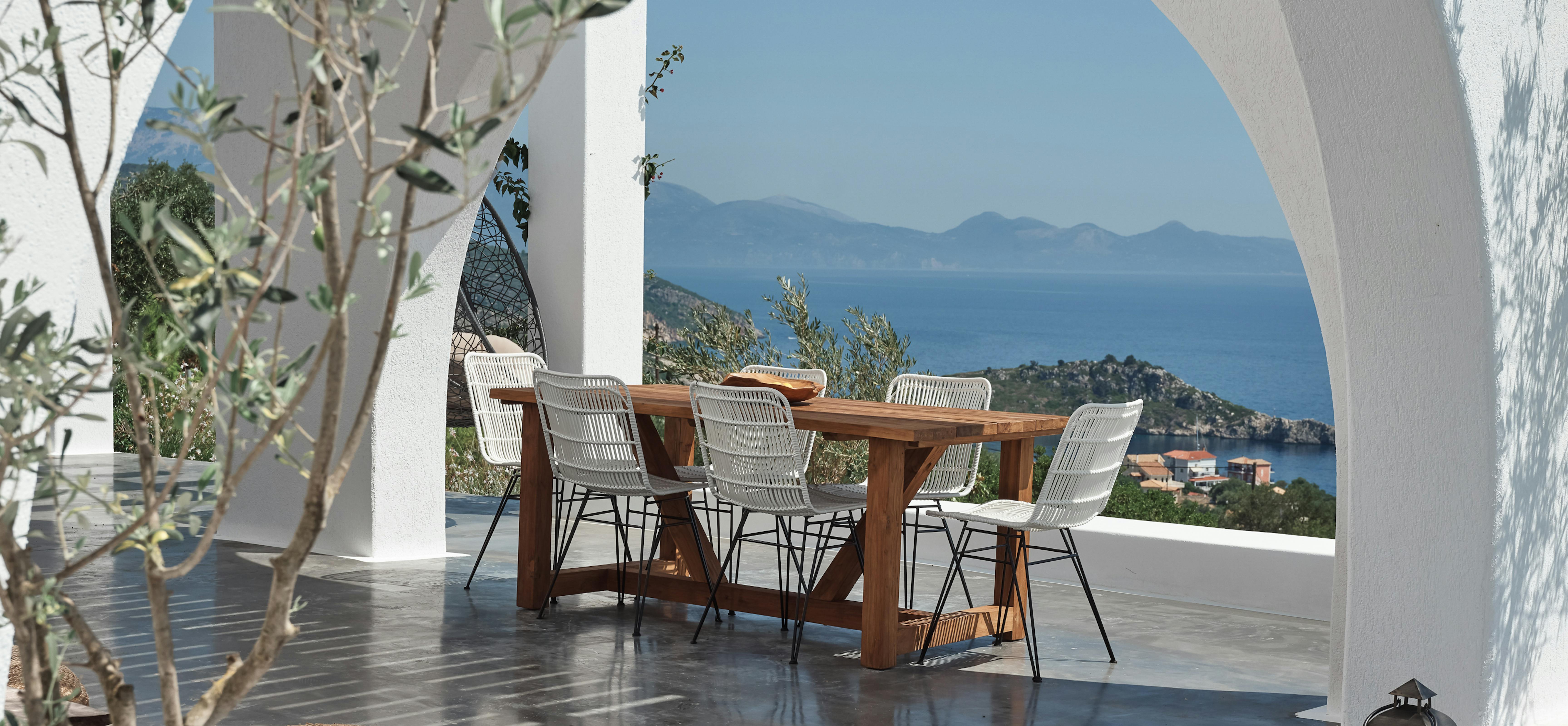 Covered dining terrace under white arched pergola frames panoramic view of Mediterranean coastline and distant mountains. White rattan chairs surround wooden table on polished concrete flooring.