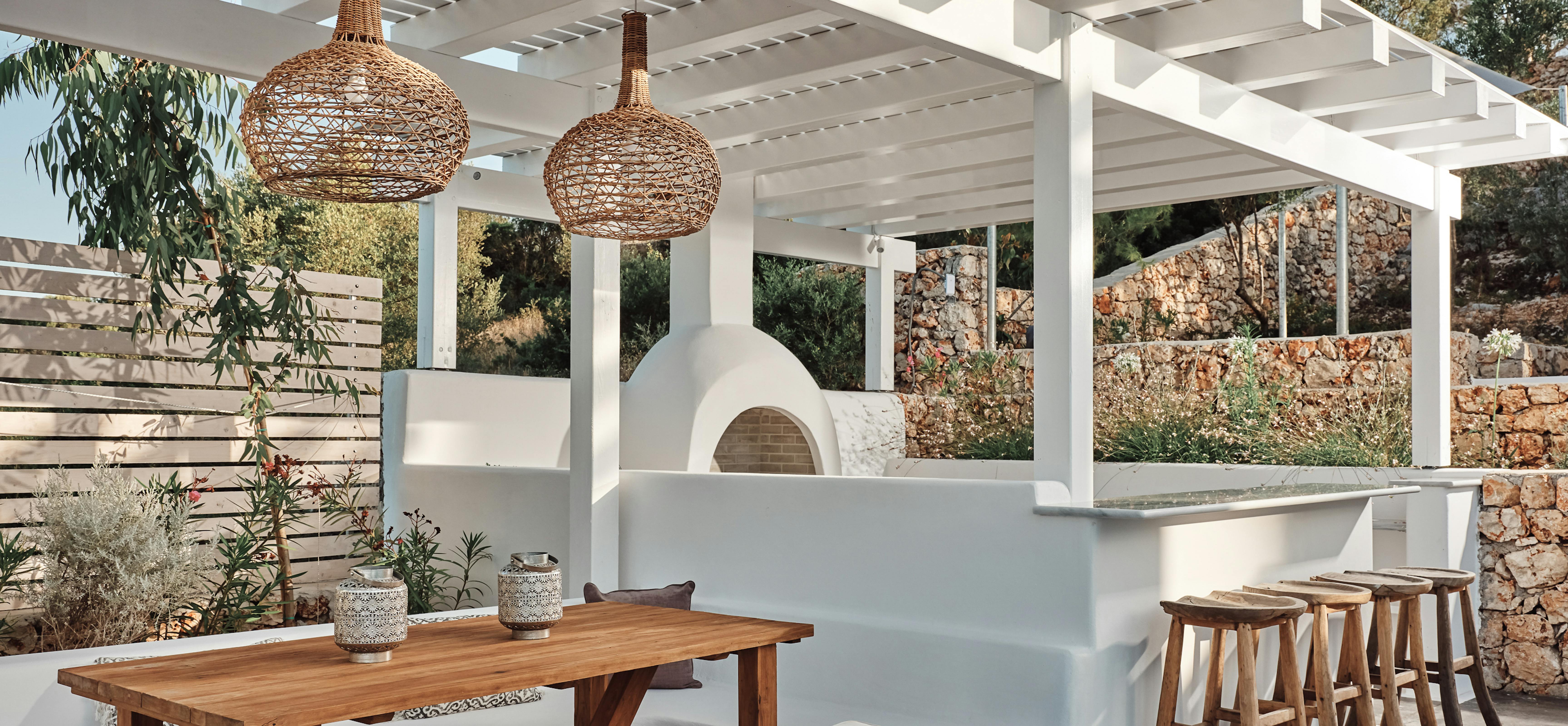 Outdoor kitchen pavilion with white pergola ceiling, wood-fired oven, and natural stone bar seating. Woven pendant lights and teak furniture create perfect entertaining space.