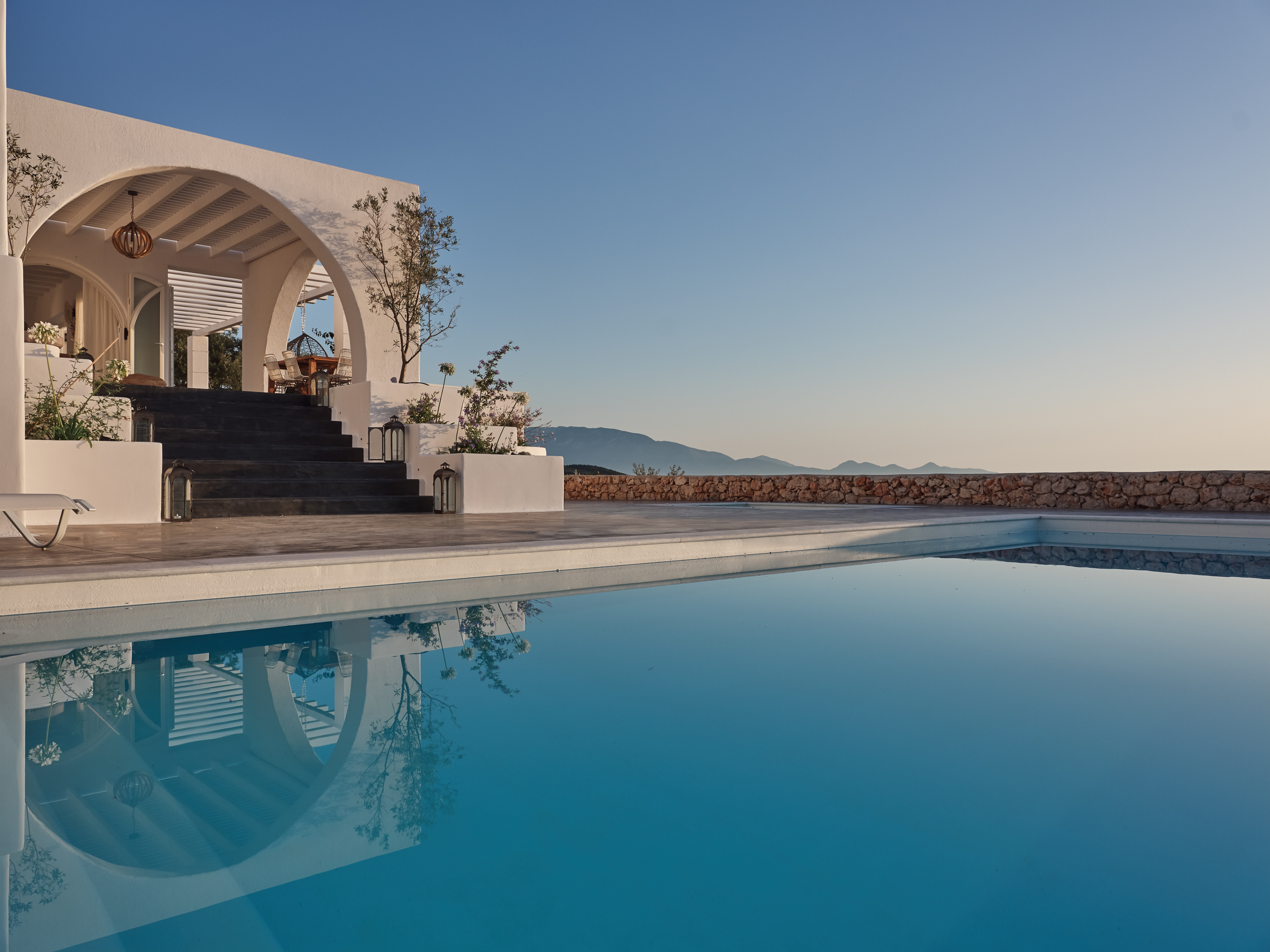 Serene infinity pool reflects white villa architecture with arched pergola structure against twilight sky. Stone retaining walls and olive trees frame the minimalist Mediterranean design.