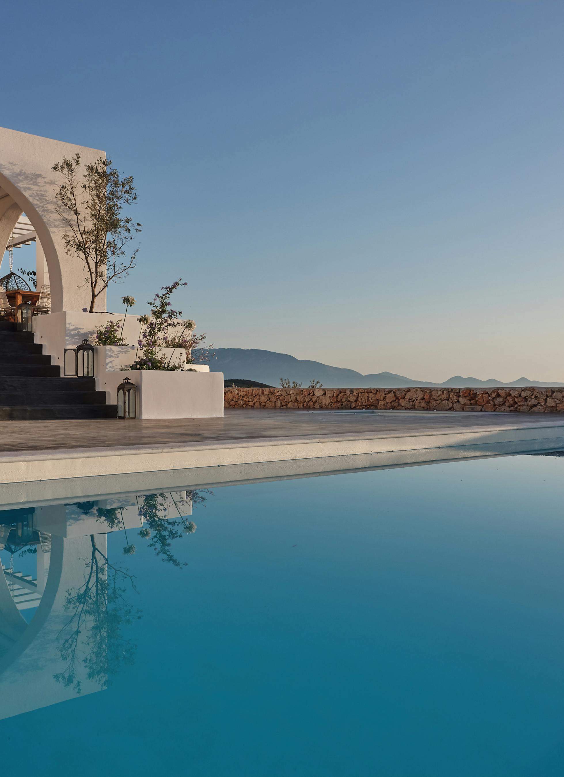 Serene infinity pool reflects white villa architecture with arched pergola structure against twilight sky. Stone retaining walls and olive trees frame the minimalist Mediterranean design.