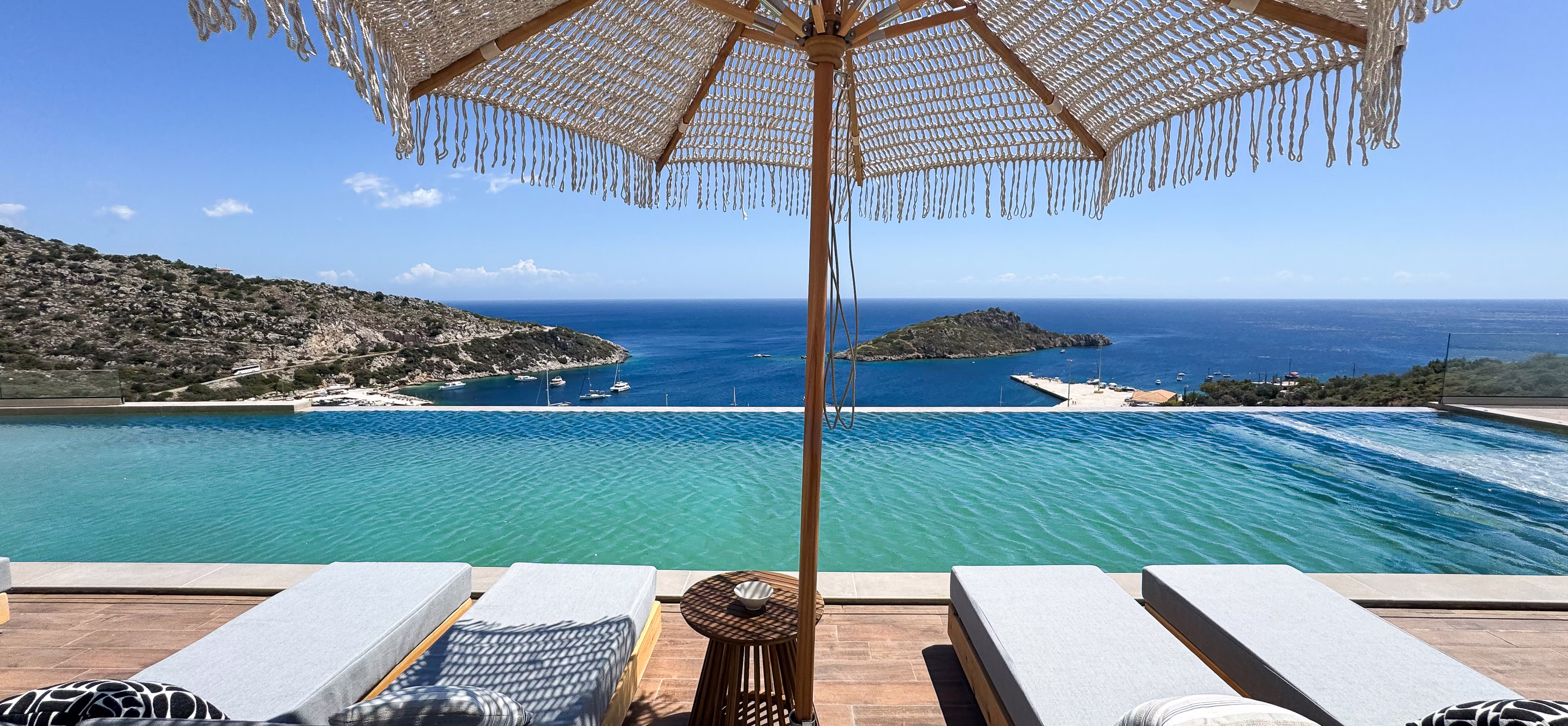 Luxury poolside lounging area with woven umbrella and teak furniture frames stunning sea views toward distant coastal islands. Grey cushioned sun beds and wicker side table create intimate outdoor relaxation space.