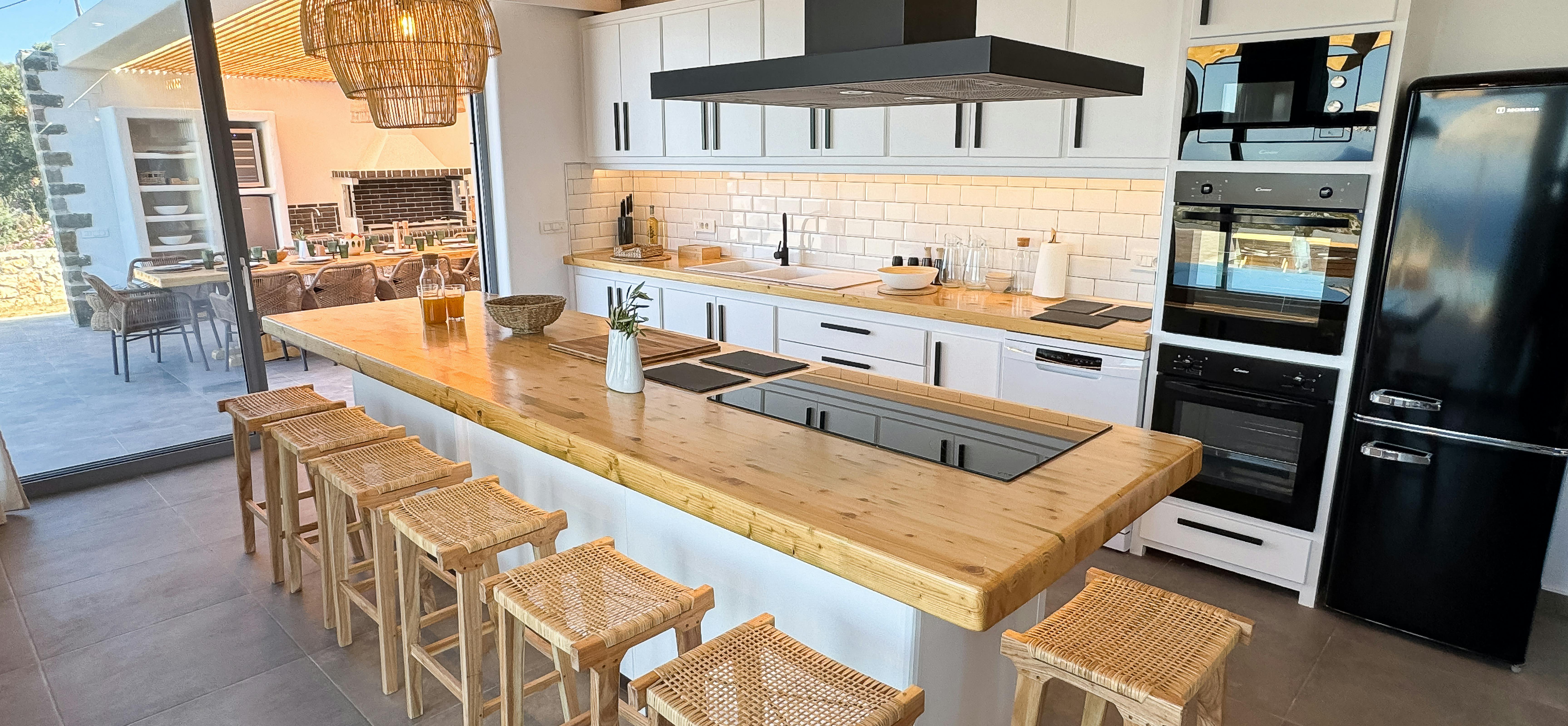 Spacious kitchen with wood beam ceiling, white cabinetry, and butcher block island seats eight on woven bar stools. Black appliances and subway tile backsplash complete this modern farmhouse-style cooking space.