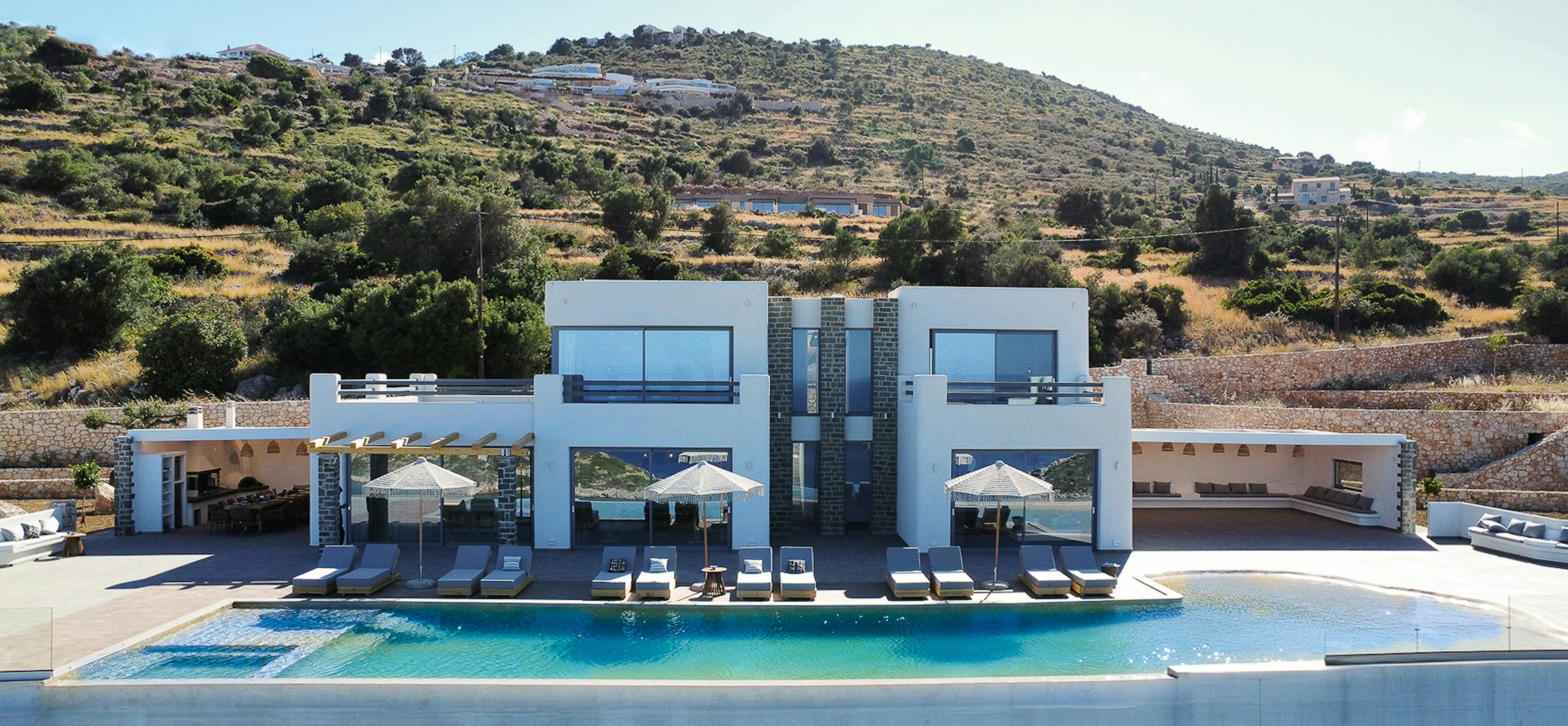 Stunning aerial view of complete villa complex showing curved infinity pool, multiple terraces, and integration with natural hillside landscape. Modern white architecture contrasts beautifully with surrounding Mediterranean countryside.