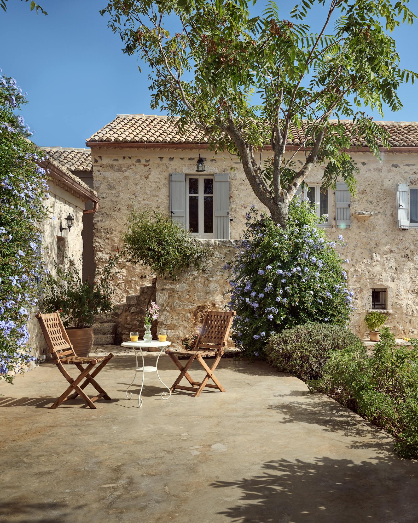Rustic stone villa with terracotta roof tiles nestled among olive trees and purple flowering shrubs. Two wooden folding chairs on flagstone terrace create peaceful garden seating area.
