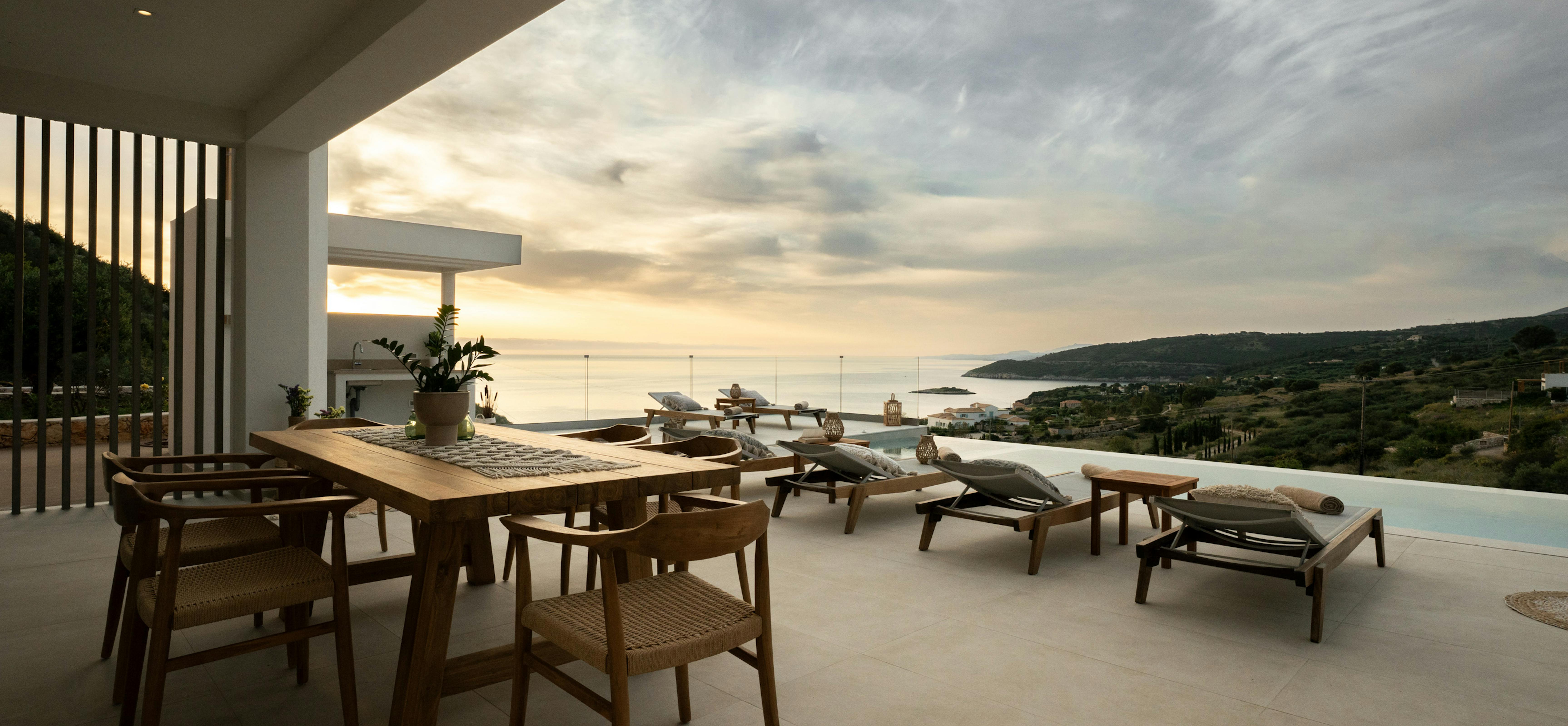 Covered terrace with wooden dining table and lounge chairs facing panoramic ocean views at golden hour. Glass railings frame the Mediterranean coastline and distant islands.