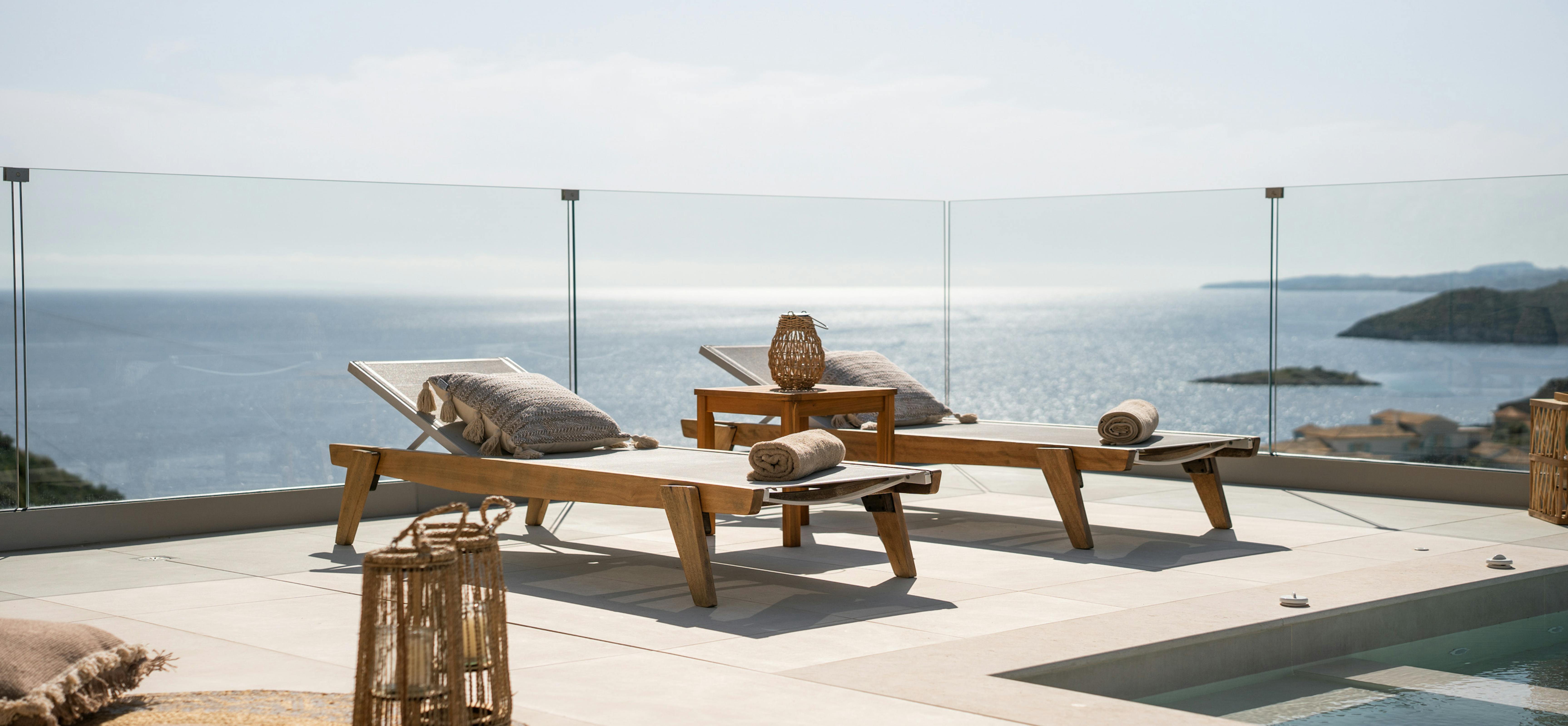 Sea-facing terrace with wooden sun loungers and glass safety railings offering unobstructed sea views. The infinity pool edge creates a seamless horizon line.
