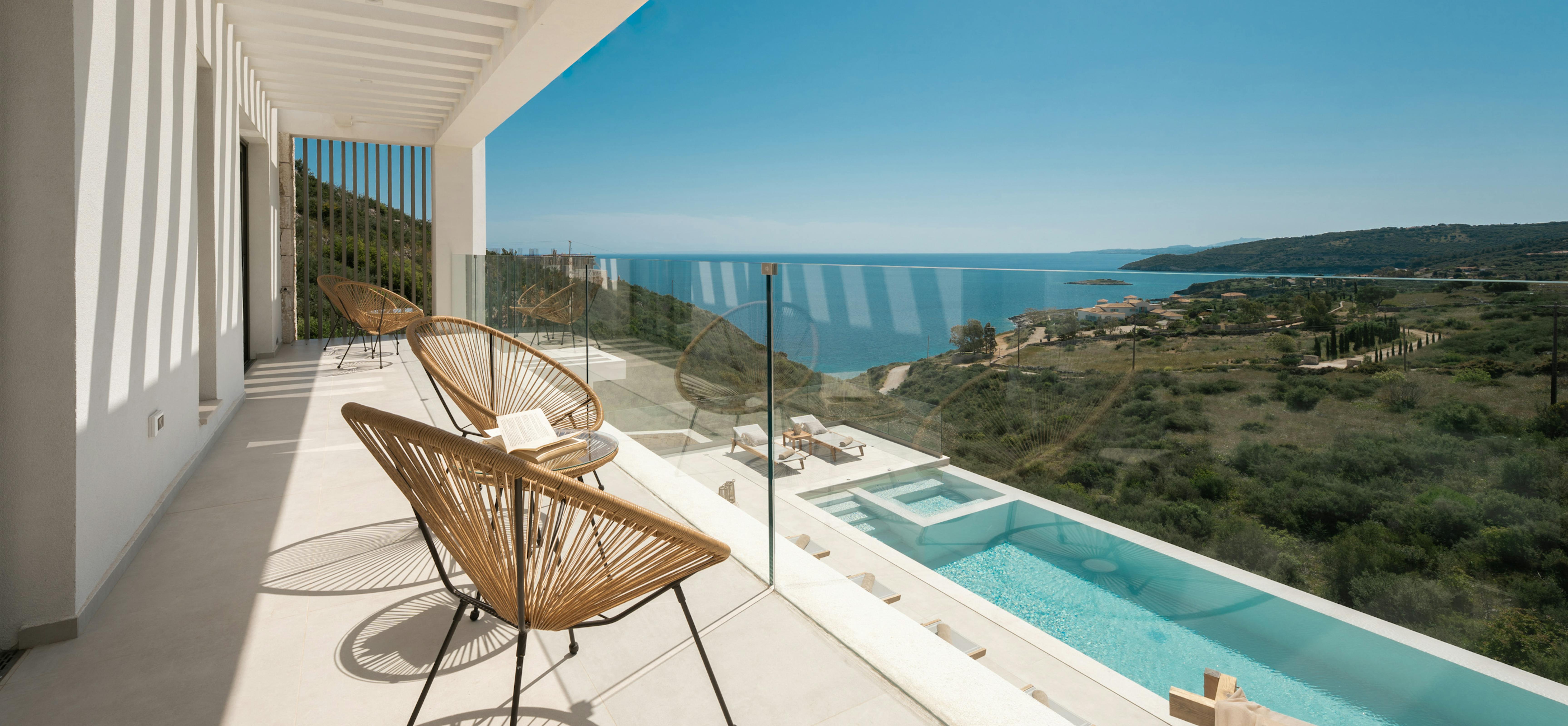 Upper-level balcony with wicker chairs overlooking pool area and distant coastline. Pergola slats create dramatic shadow patterns on the neutral stone flooring.