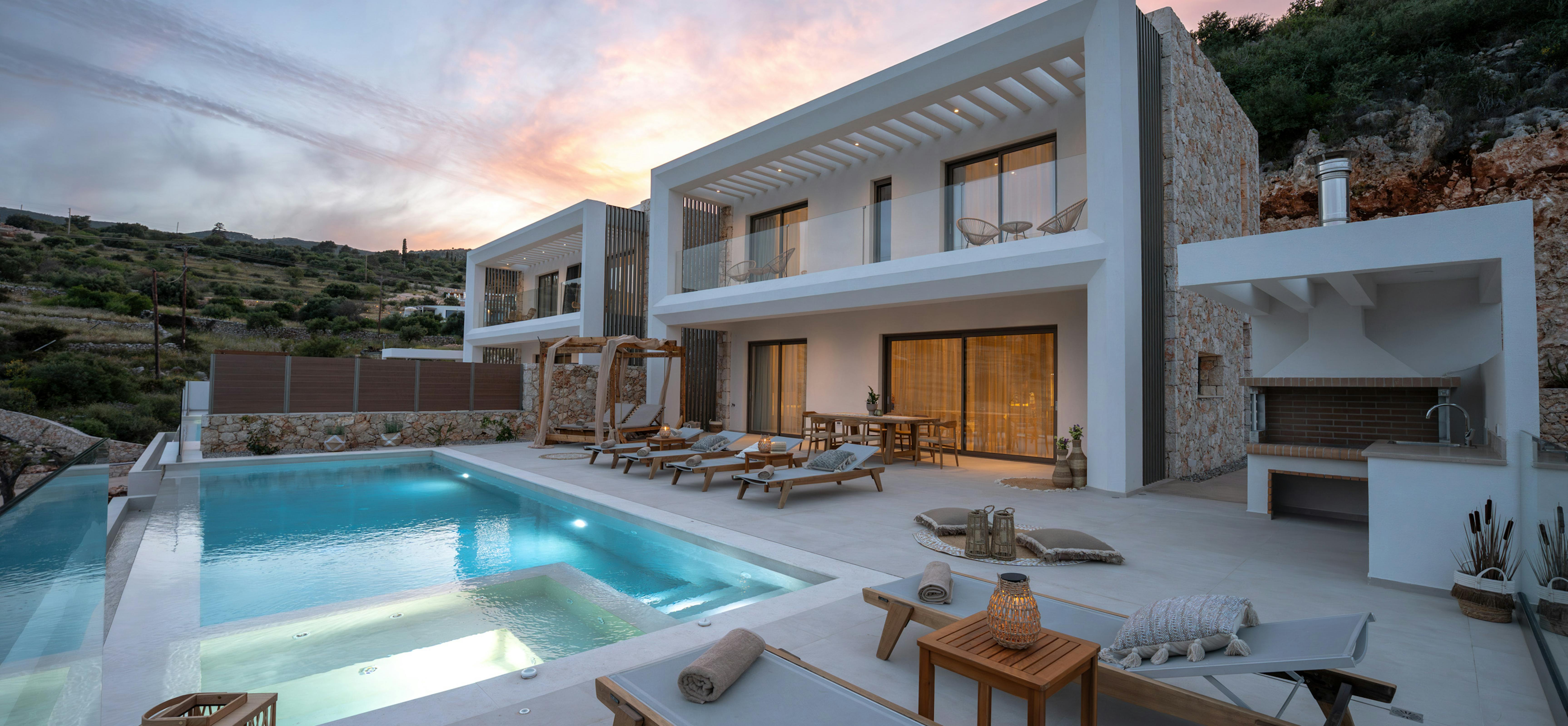 Exterior view of villa at dusk with illuminated pool area and warm interior lighting. The modern architecture contrasts beautifully against the natural hillside setting.