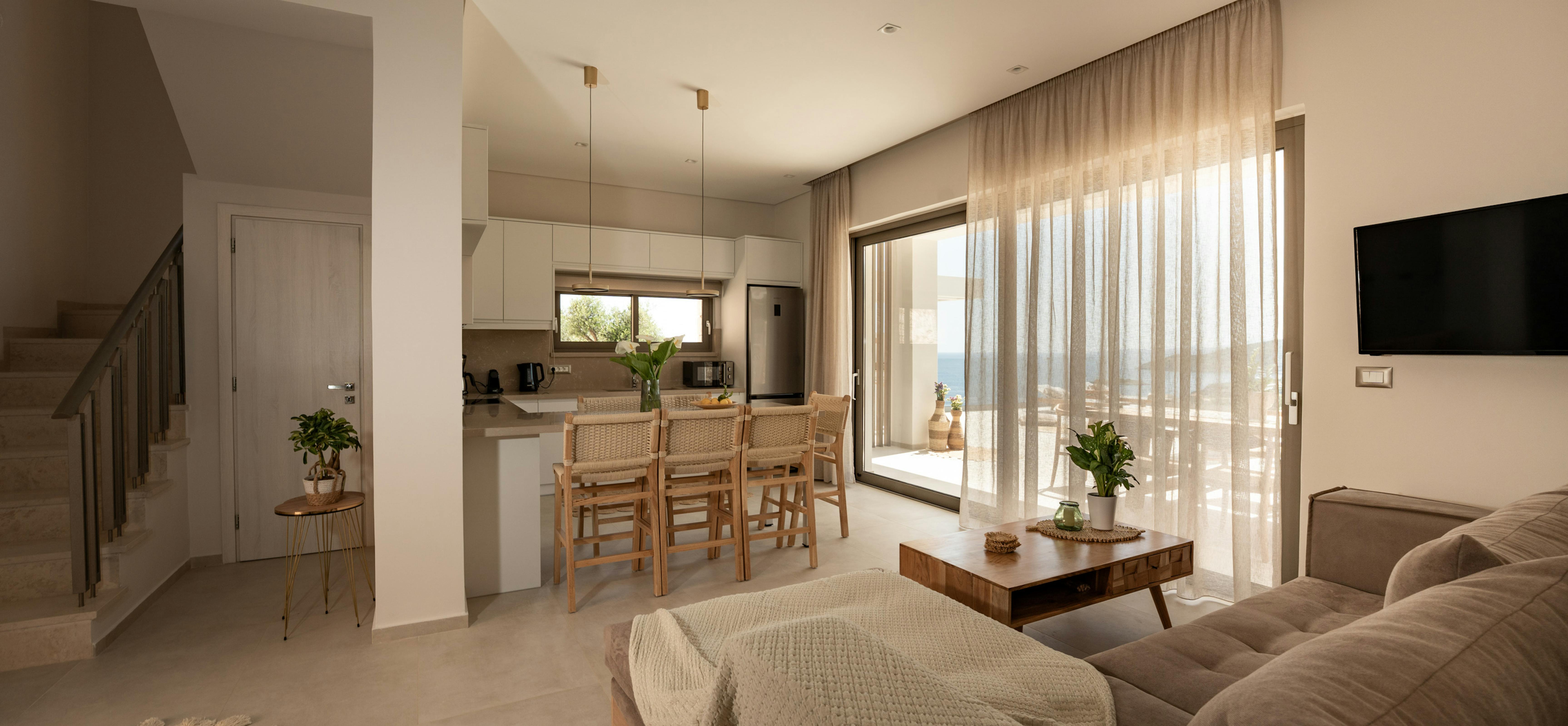Open-plan living area with white kitchen, woven bar stools, and sectional sofa overlooking a terrace. Stairs lead to upper level in this contemporary Mediterranean home.