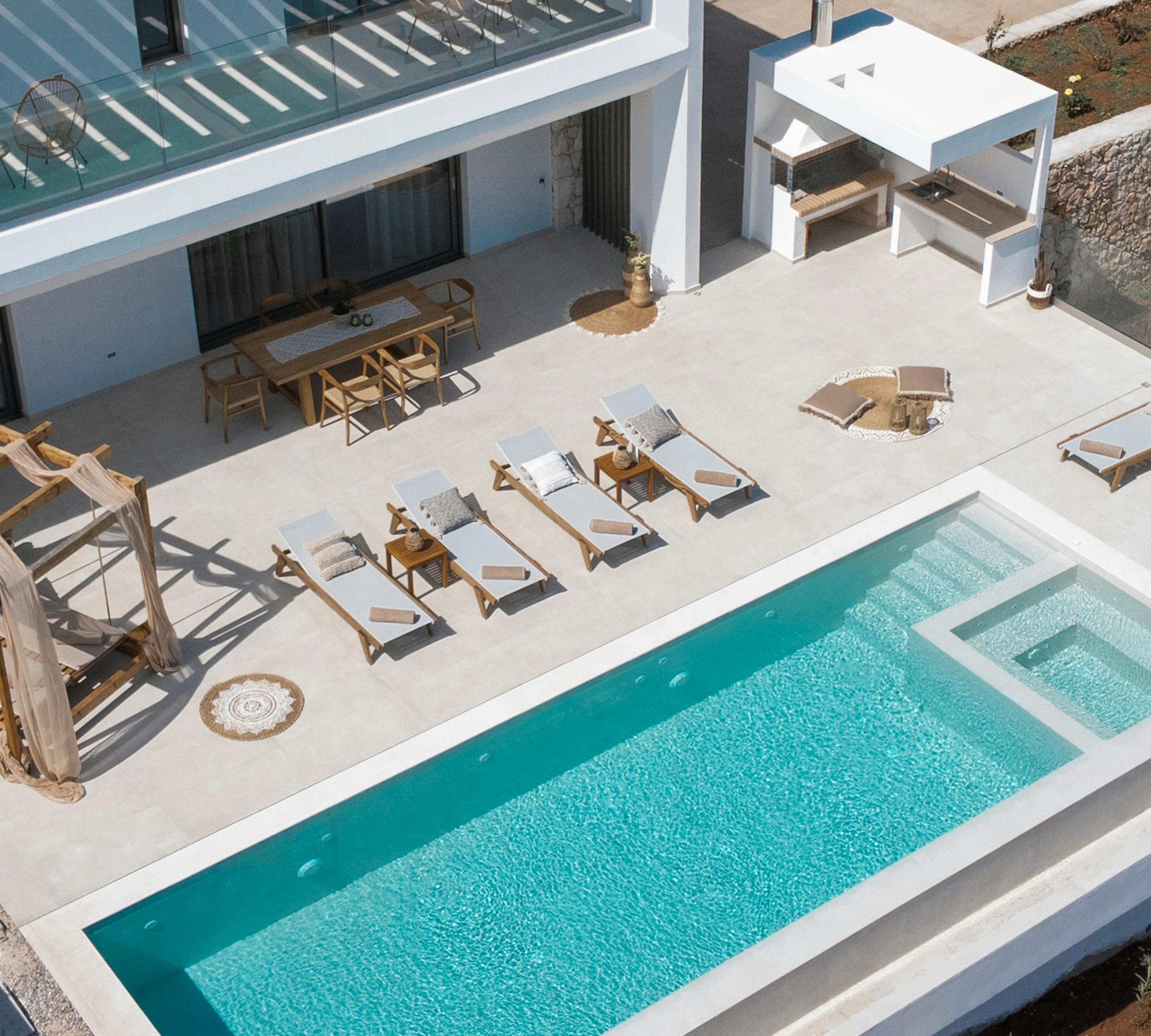 Aerial view of modern villa's pool deck with rectangular infinity pool, wooden sun loungers, and geometric outdoor furniture arrangement. Clean white architecture contrasts with surrounding landscape.